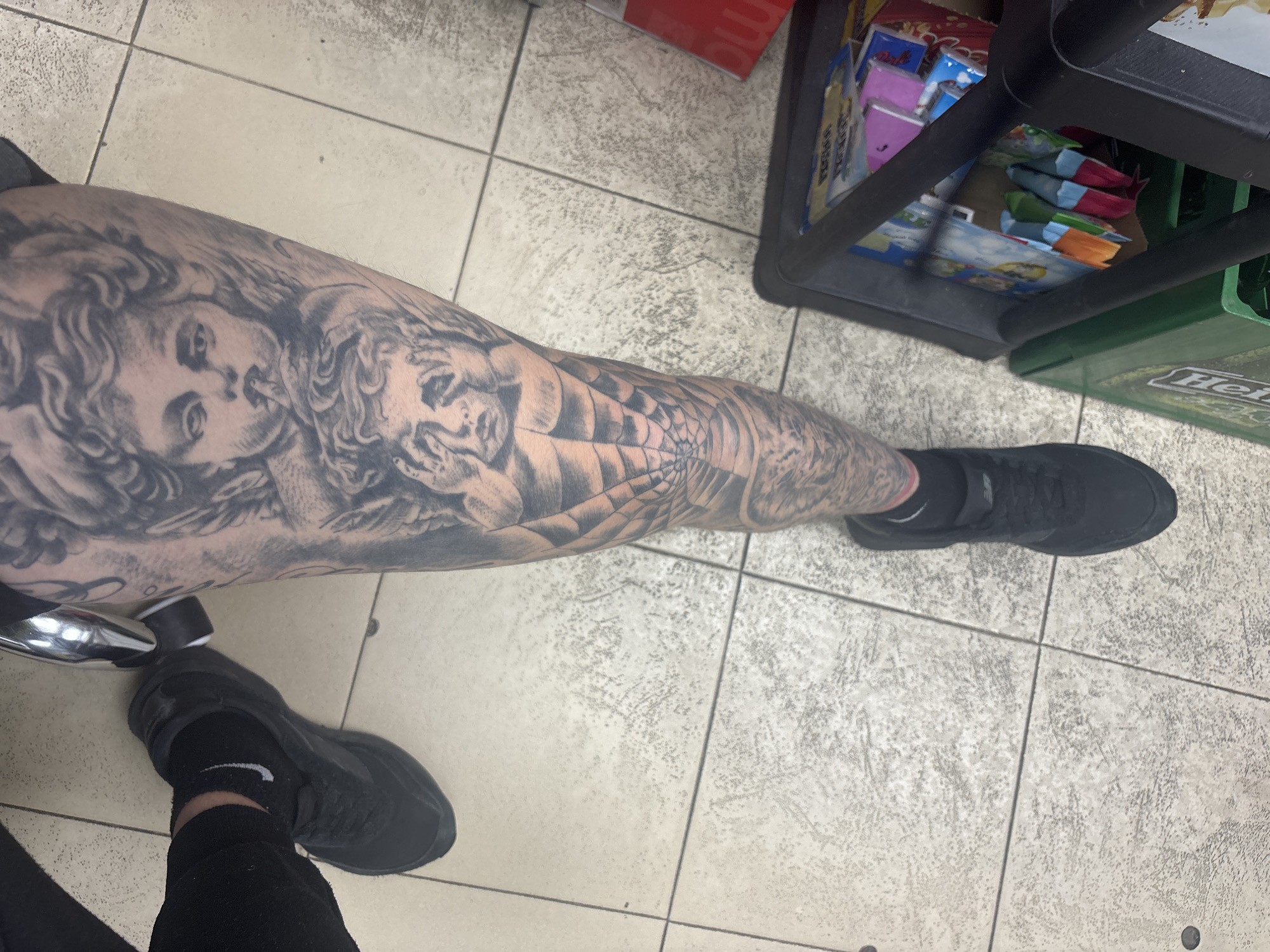 The image shows a person's tattooed left leg and foot. The tattoos depict two angelic figures and a spiderweb pattern. The person is wearing black socks and black sneakers. They are standing on a tiled floor, next to a black plastic shopping cart or shelf unit containing various packaged goods. A green Heineken beer crate is partially visible behind the cart. The location is likely indoors, possibly a store. There is no explicit indicator of the city of Sofia, Bulgaria.
