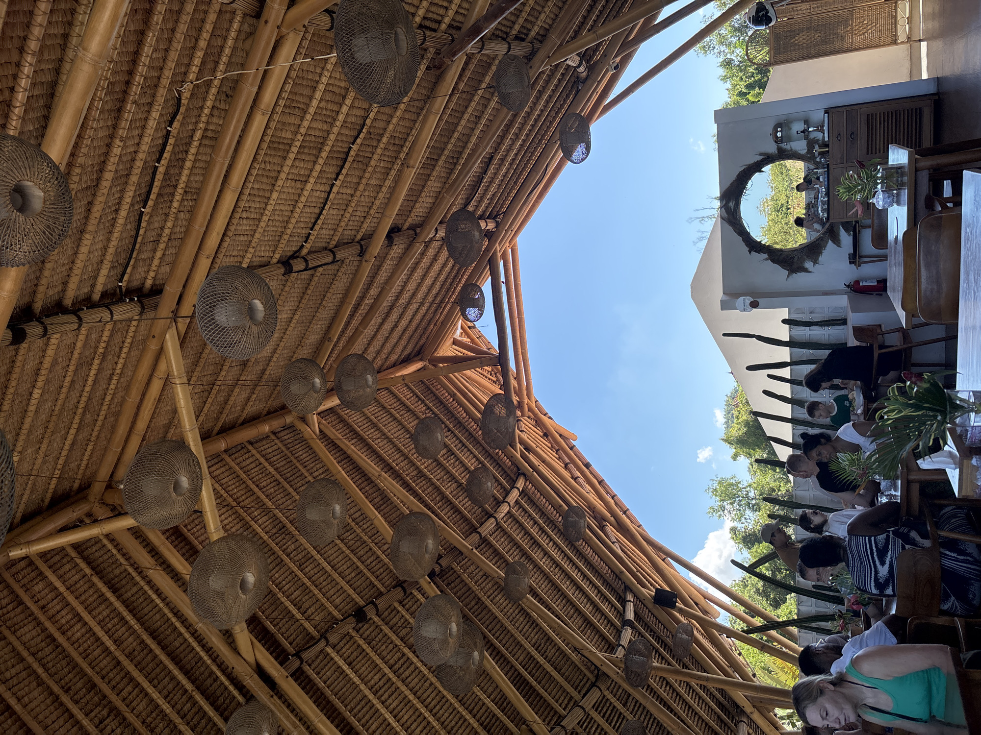 The image shows an indoor dining area in Kuta Selatan, Indonesia. The roof is made of bamboo and thatched material with round decorative lamps suspended from it. A gap in the roof reveals a clear blue sky. A group of people are seated at tables on the right, with plants visible between them. In the background, there is a white wall with a round mirror decorated with straw. A wooden cabinet sits against the wall.