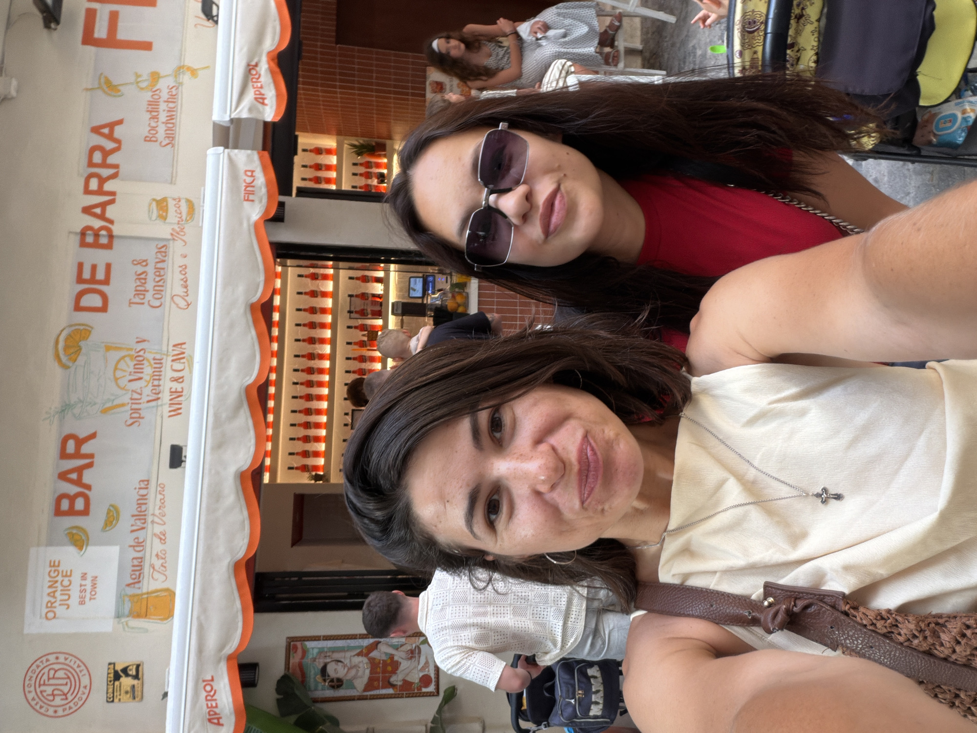 Two women are taking a selfie in front of a bar. The woman in the foreground on the left has dark brown hair and is wearing a cream-colored top with a brown leather strap bag over her shoulder. She is wearing a silver necklace with a cross pendant. The woman to her right has dark brown hair and is wearing sunglasses and a red t-shirt. Behind them, the bar has an orange and white awning with "APEROL" written on it. The bar has a display of red bottles and some orange juice. A man in a white patterned shirt and light shorts is standing in the background, looking away from the camera, and pushing a stroller. Another woman is sitting in the background on the right. The scene appears to be outdoors, possibly on a street or promenade.