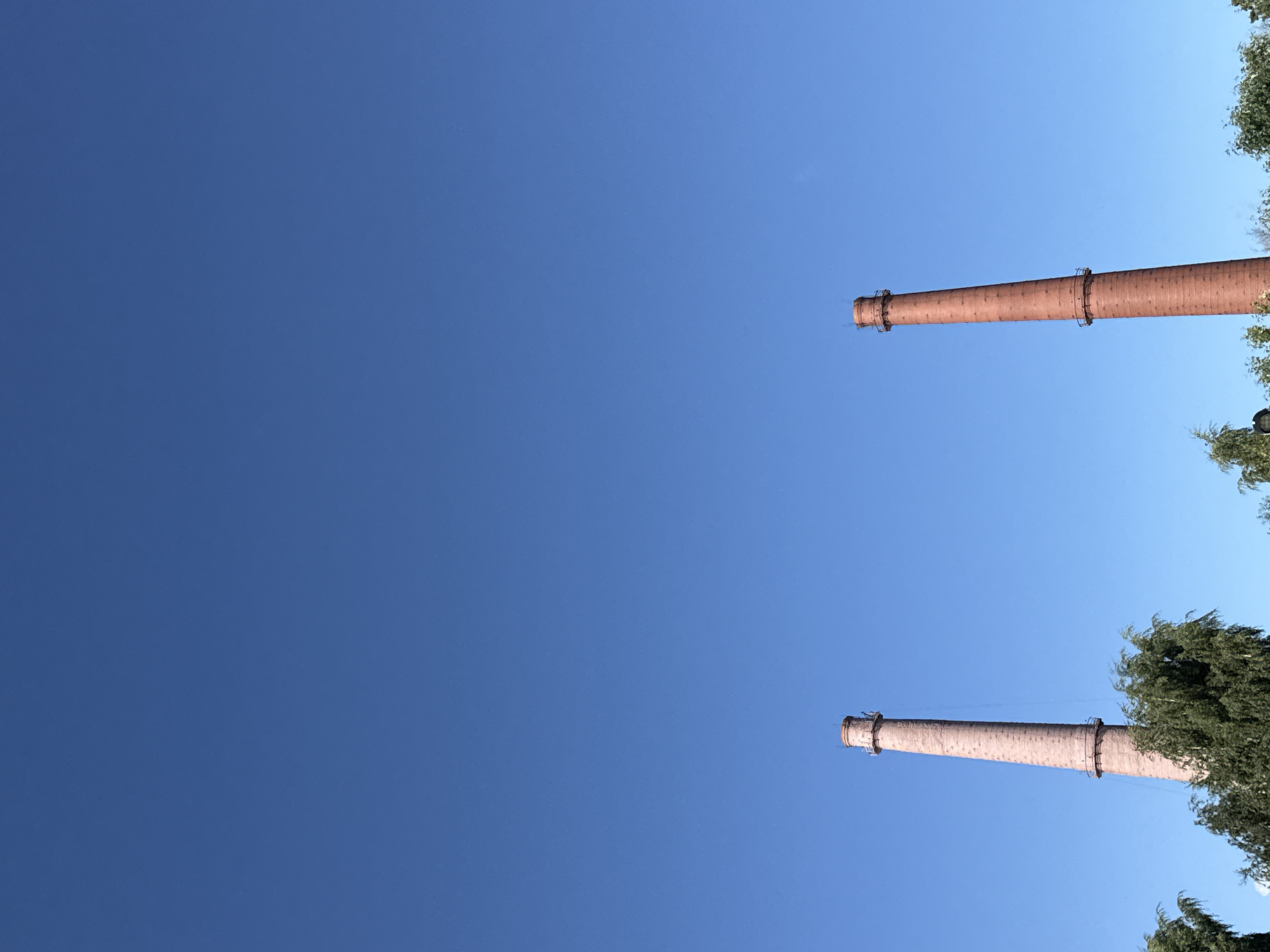 The image displays two tall brick smokestacks against a clear blue sky. The smokestack on the left is lighter in color, appearing to be made of a pinkish-red brick, and is partially obscured by green foliage at its base. The smokestack on the right is a deeper red brick and stands against the sky with no obstructions. Both smokestacks have visible bands and railings around their upper sections. The scene is devoid of people and activity, presenting a stark, industrial landscape. It is possible these smokestacks are located in Kronstadt, Russia, a city known for its historical maritime and industrial infrastructure.