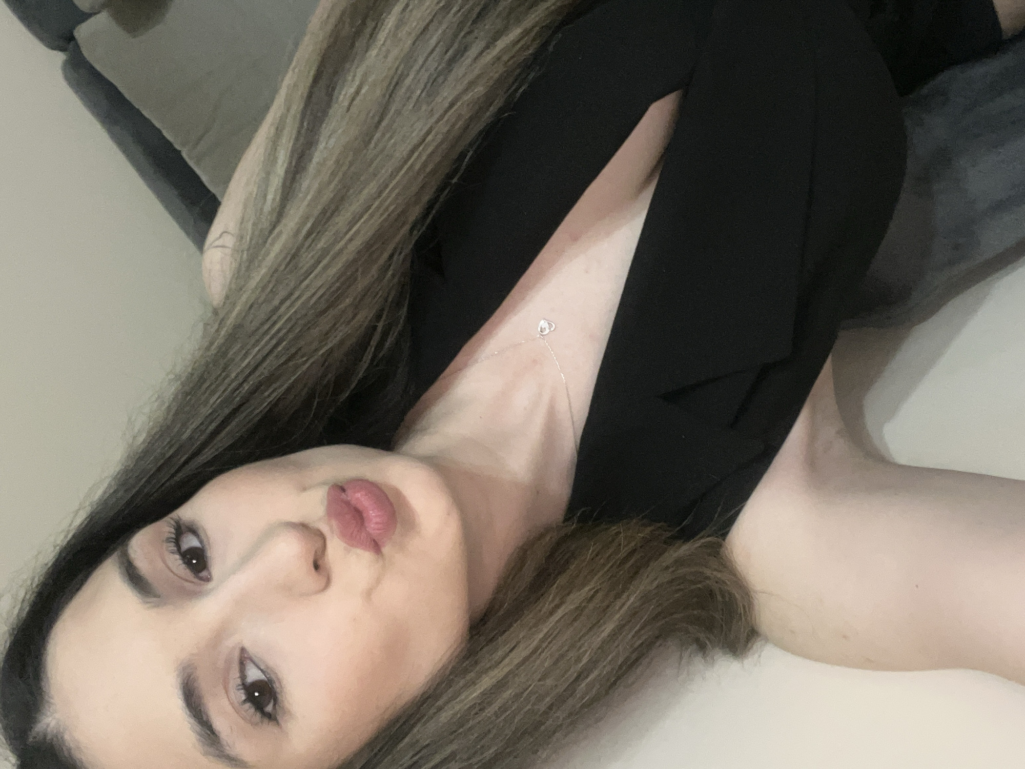 The image is a selfie of a young woman with long brown hair, wearing a black top and a delicate silver necklace. She is posing with her lips puckered, looking towards the camera. The background is softly blurred, showing a neutral-colored wall and a hint of a piece of furniture. There is a tattoo visible on her left shoulder. The setting appears to be indoors, possibly a home or a hotel room. There is no information available to determine the location in Plovdiv, Bulgaria, or any specific details about the environment beyond what is visible.