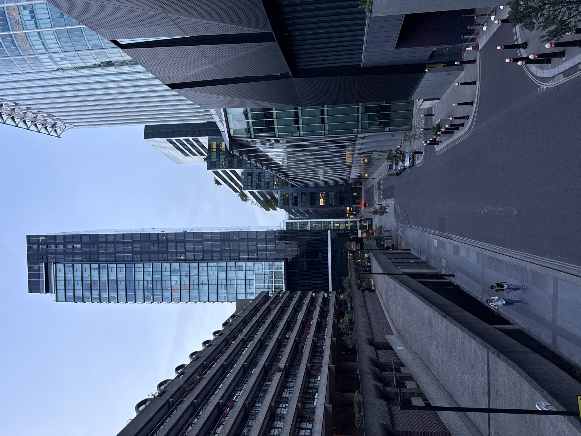 The image depicts an urban scene in London, United Kingdom, showcasing a street lined with modern and brutalist architectural styles. Several tall buildings, constructed with glass and concrete, dominate the skyline. The image captures a narrow roadway and a sidewalk where two people are walking. The overall atmosphere is somewhat muted, possibly taken during early morning or late evening, with the sky visible above.