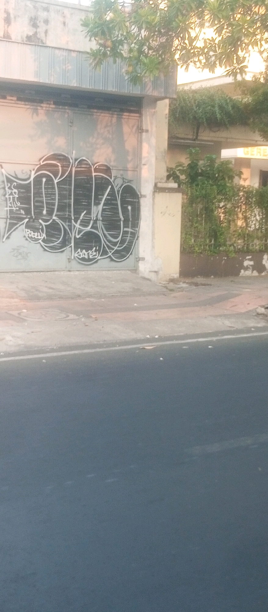 The image shows a gray, corrugated metal gate on a building in Kediri City, Indonesia. The gate is covered in black graffiti in a bubble letter style. To the right of the gate is a light yellow concrete pillar and a green metal fence with dense foliage behind it. A white sign with yellow lettering is visible above the fence. In front of the gate is a gray concrete sidewalk. The street in front of the sidewalk is paved with asphalt and has a white line marking. The scene is illuminated by natural light, suggesting it was taken during the daytime. There are no people visible in the image.