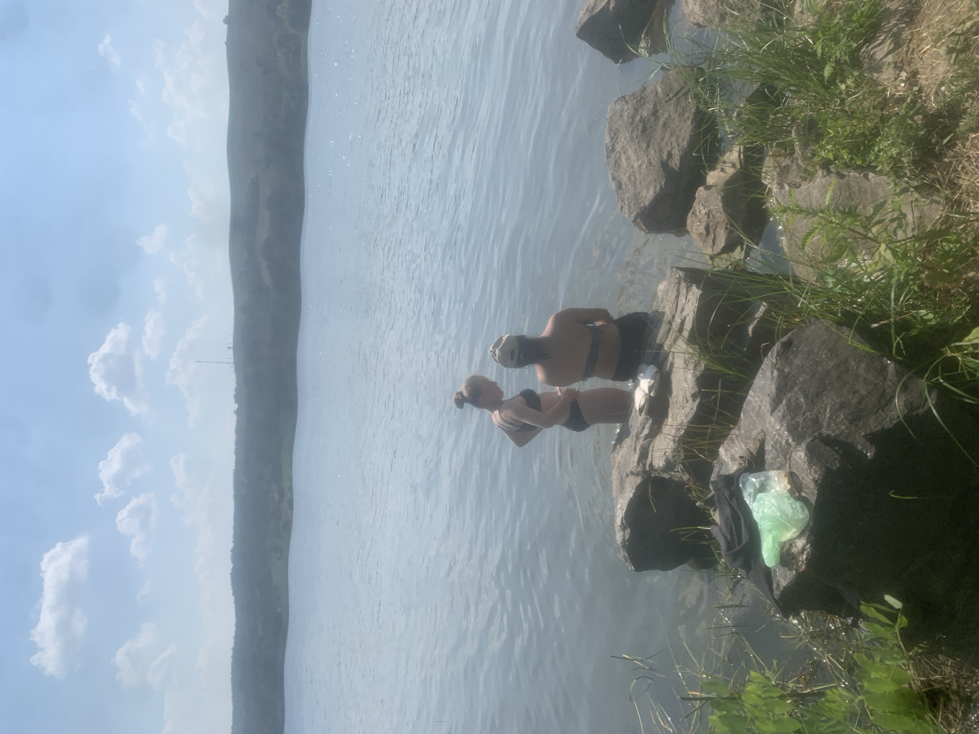 Two young women are wading in a body of water near the shore, which is lined with large rocks and vegetation. The woman closer to the shore is wearing a black bikini and has her hair tied up in a bun. She is looking away from the camera and appears to be interacting with the other woman. The second woman, partially submerged in the water, is also wearing a black bikini. She is facing the camera and has dark hair. The water is calm with ripples on its surface, reflecting the light blue sky and scattered white clouds. In the background, a distant, hazy shoreline with a gradual slope can be seen. The sun appears to be high in the sky, suggesting it is daytime, and the weather seems to be warm and pleasant. On the rocks near the shore, there are some discarded items, including a black piece of clothing and a bright green plastic bag, indicating a casual outing or recreation by the water. The overall scene depicts a relaxed summer day at a natural waterfront location.