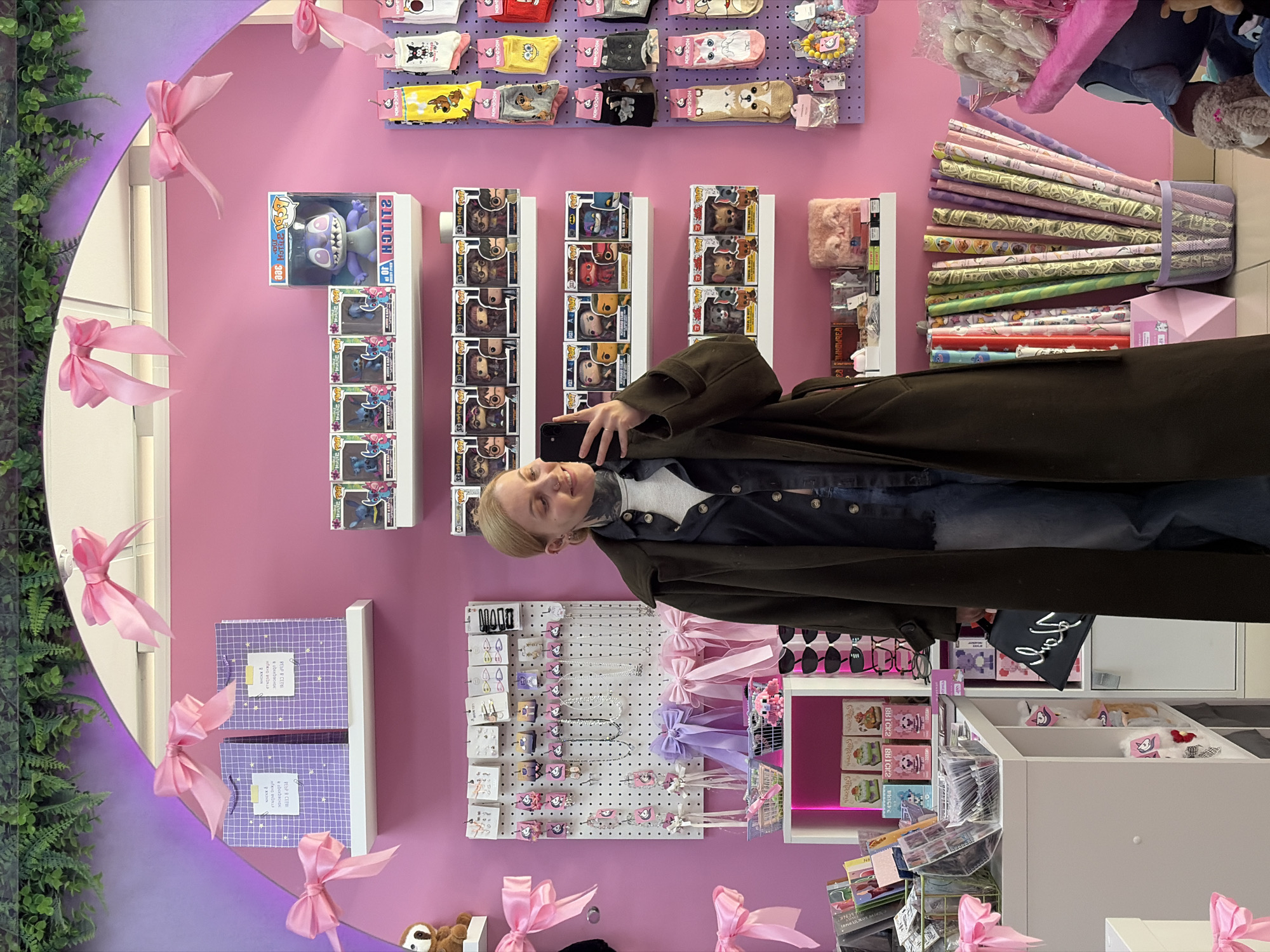 A blond woman in a green coat takes a selfie in a pink-walled shop, Chaikovsky, Russia. There are shelves of Funko Pop figurines, socks, and jewelry on the wall. Wrapped rolls of colorful paper are stacked high on a shelf to the right. A mirror with pink ribbons and faux plants surrounds the left side of the frame. Boxes and miscellaneous products are stacked on shelves below.