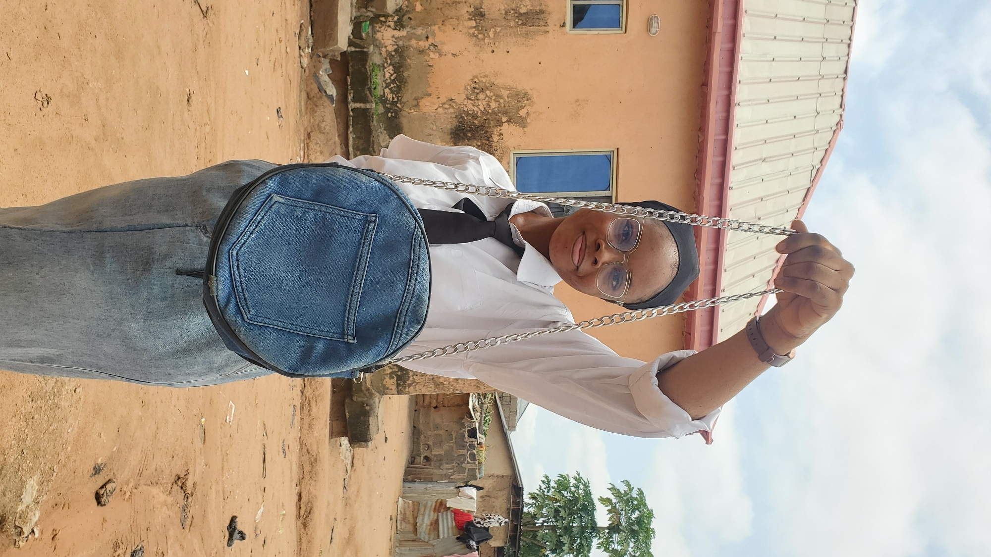 A young woman stands outdoors, holding a round denim handbag by its silver chain strap. She is wearing a white collared shirt with a black tie, a black headscarf, and glasses. Her attire suggests a school uniform or a formal ensemble. She is looking towards the camera with a slight smile. The background features a peach-colored building with visible architectural details, windows, and a corrugated metal roof. The ground is unpaved and sandy, with scattered debris. To the right of the building, there are more structures made of brick and corrugated metal, with laundry hanging to dry. The sky above is partly cloudy. The overall impression is that of a residential area in a town or city, possibly in Nigeria, given the architectural style and the presence of laundry. The scene is lit by natural daylight.