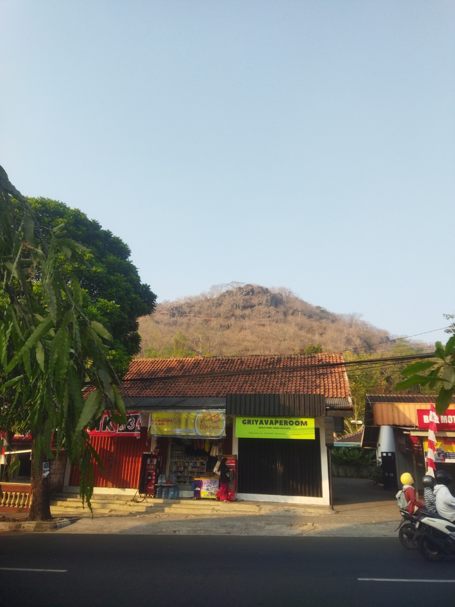 The image captures a street scene in Wonogiri, Indonesia, featuring a collection of shops and a prominent, dry, tree-covered hillside in the background. In the foreground, a paved road runs horizontally across the bottom of the frame. To the left, a large tree with lush green foliage partially obscures the view. Adjacent to the tree, several shops are visible. One shop has a red rolling door with white text that appears to be "IK34". Another shop displays a yellow banner with green text reading "GRIYAVAPEROOM" and a tagline below it. Further to the right, a shop with a red sign and a white and red striped flag can be seen, labeled "BO MOT...". Two individuals are on a motorcycle in front of this shop, one wearing a yellow helmet and the other a dark helmet. Another person, dressed in red, is standing in front of the shop with the yellow banner. The shops have tiled roofs and appear to be single-story structures. The hillside in the background is largely barren, with dry trees and rocky outcrops, indicating a dry season. The sky is clear and pale blue. The overall lighting suggests it is daytime, possibly late afternoon due to the long shadows cast.