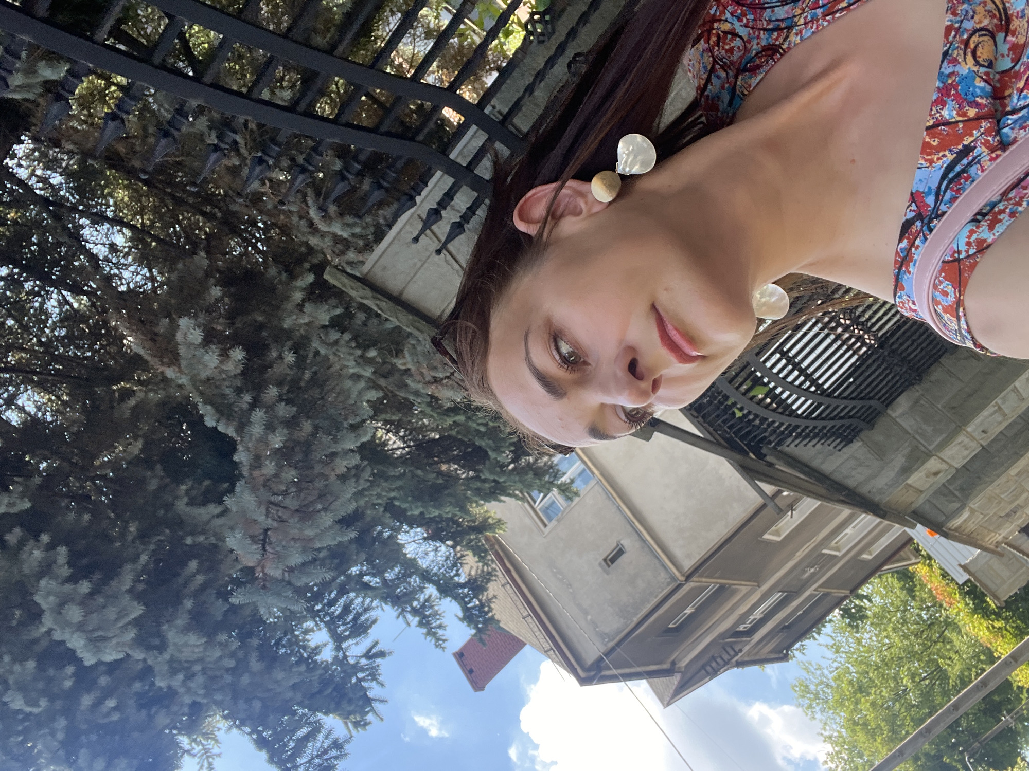 The image is a close-up, sideways selfie of a young woman. She has long, dark brown hair and is wearing a colorful, patterned top. She has two large, round earrings in her ears, one light brown and one pearlescent. Her eyes are looking towards the camera, and she has a slight smile on her lips. The background shows a residential area with a house and trees. A black metal fence is visible in the upper part of the image, and the sky is blue with some white clouds. The lighting suggests it is daytime, possibly sunny. The setting appears to be outdoors, possibly a street or backyard. The woman's expression seems relaxed and happy.