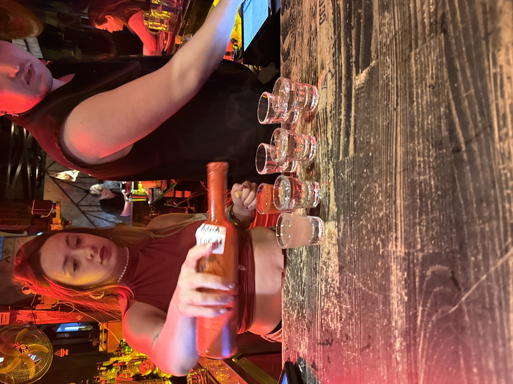 The photograph captures a scene within a bar environment, possibly located in Batumi, Georgia. A blonde woman in a dark red top stands behind the bar, pouring liquid from a bottle labeled "Adja Sugar" into a row of clear shot glasses lined up on a wooden bar top. There are six shot glasses, the last one being partially filled. Another person is seen standing behind the counter next to her. Other people are visible in the background, along with an array of bottles typically found in a bar setting. The lighting is dim and reddish, suggesting an evening or nighttime setting. A vintage fan is mounted nearby.
