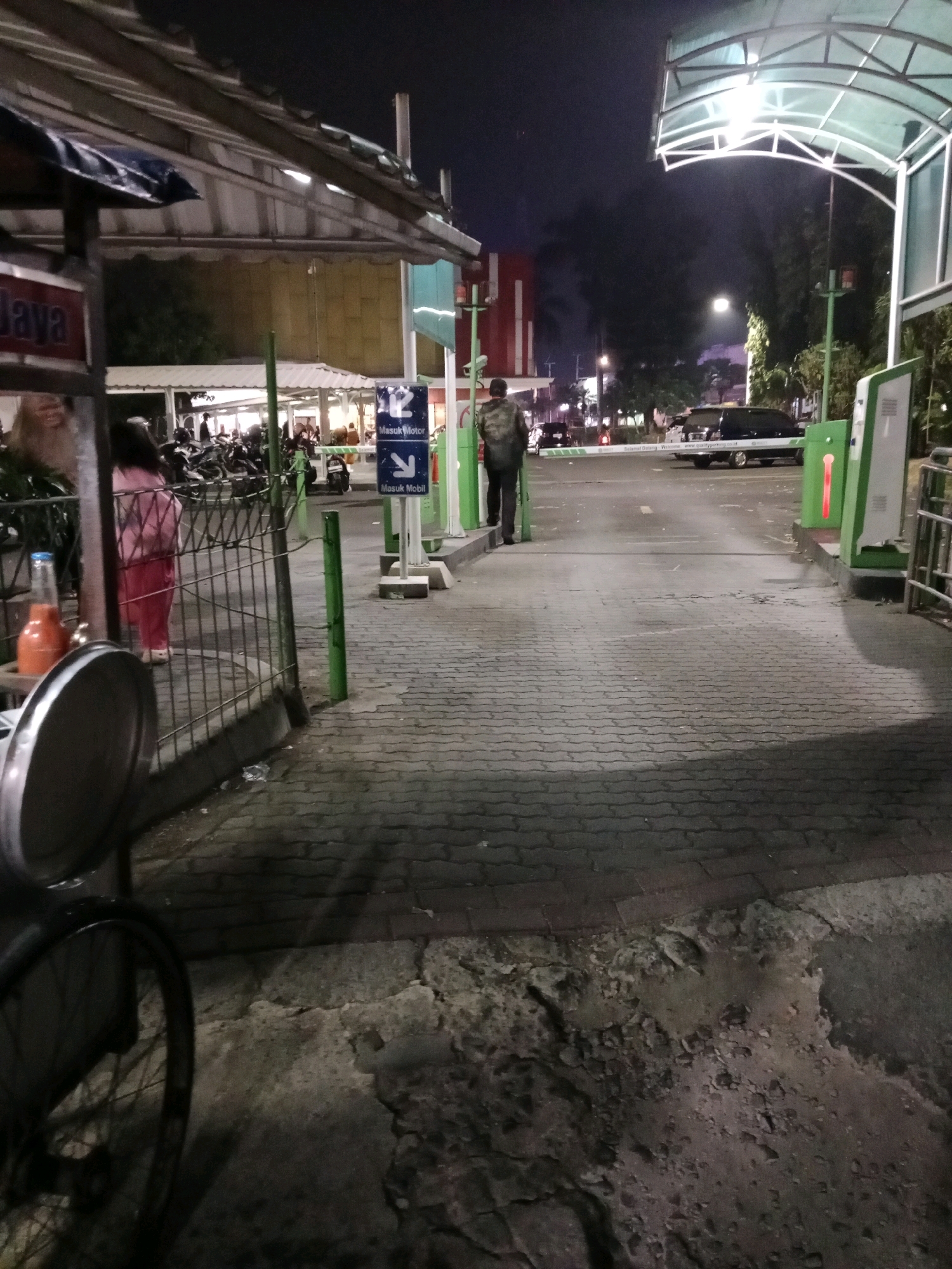 The image depicts a nighttime scene at a parking facility, likely an entrance or exit, in Cianjur, Indonesia. A paved pathway, made of interlocking bricks, leads towards a barrier arm. On the right, a green and white automated payment or access machine is visible, illuminated by a red light. A person wearing a dark jacket and pants walks away from the camera towards the barrier. In the background, several motorcycles are parked, and a dark-colored minivan is visible. Above the pathway, a white canopy structure with integrated lighting provides illumination. A sign with blue and white markings, indicating "Masuk Motor" (Motorcycle Entry) and "Masuk Mobil" (Car Entry) with corresponding arrows, is mounted on a pole. To the left, a metal fence encloses an area where a person in a pink shirt and pants stands. A bicycle with a chrome-colored wheel is partially visible in the foreground on the left. The overall environment suggests a well-lit but somewhat worn urban parking area.