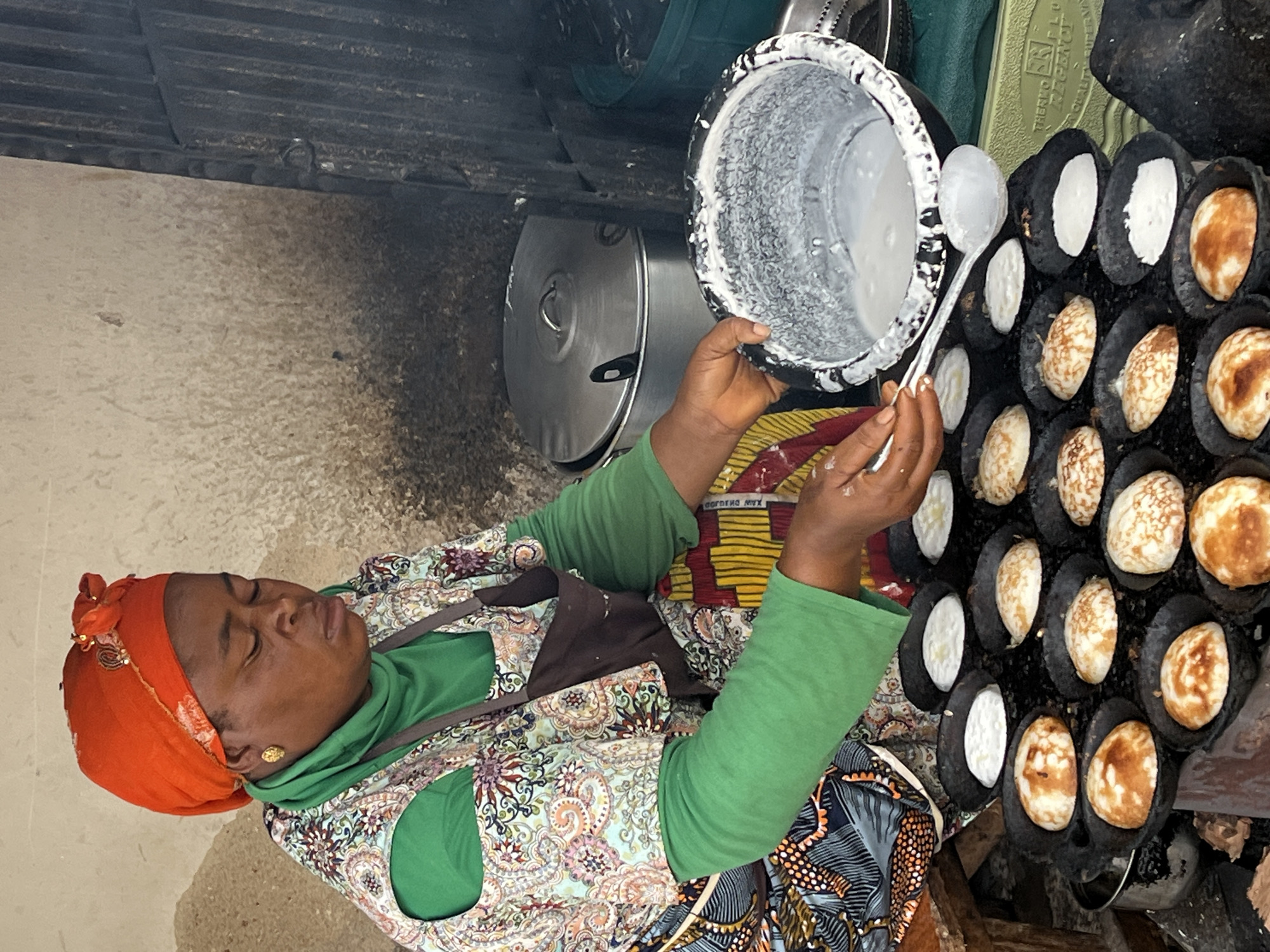 A woman wearing an orange headscarf, a green hooded shirt, and a patterned dress is preparing food in an outdoor setting, likely a marketplace, in Jos, Nigeria. She is actively cooking what appear to be small, round pancakes or fritters in a specialized pan with multiple molds. The pan is dark and set over a heat source, with some of the cooked items showing a golden-brown color, while others are still white, indicating they are at different stages of cooking.
In her right hand, she holds a ladle filled with a white batter, which she is in the process of pouring into one of the empty molds in the pan. Next to her, a large, lidded metal pot suggests she is preparing a larger quantity of food or has other cooking ingredients nearby. The background shows a textured wall and some wooden elements, typical of an outdoor cooking area. The woman's eyes are closed, and her expression appears focused and perhaps slightly strained, suggesting concentration on her task. The time of day is indeterminate, but the lighting suggests it could be daytime. No specific weather conditions are evident.
