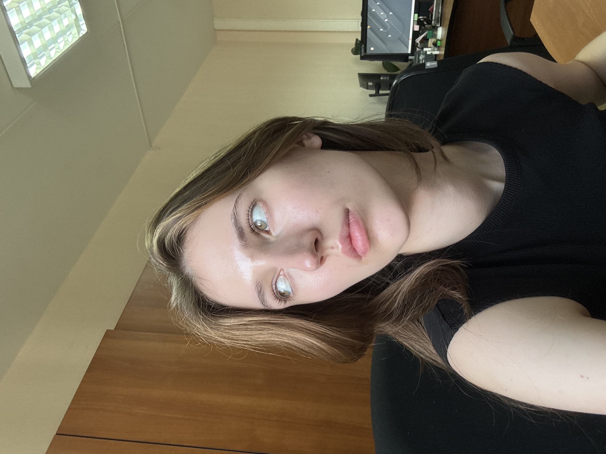 The image shows a young woman with light brown hair and green eyes, looking to her left. She is wearing a black sleeveless top and appears to be indoors, possibly in an office or residential setting. The background includes a wooden cabinet, a ceiling light fixture, and a desk with computer equipment. The image does not provide any specific information about the location within the city of Perm, Russia.