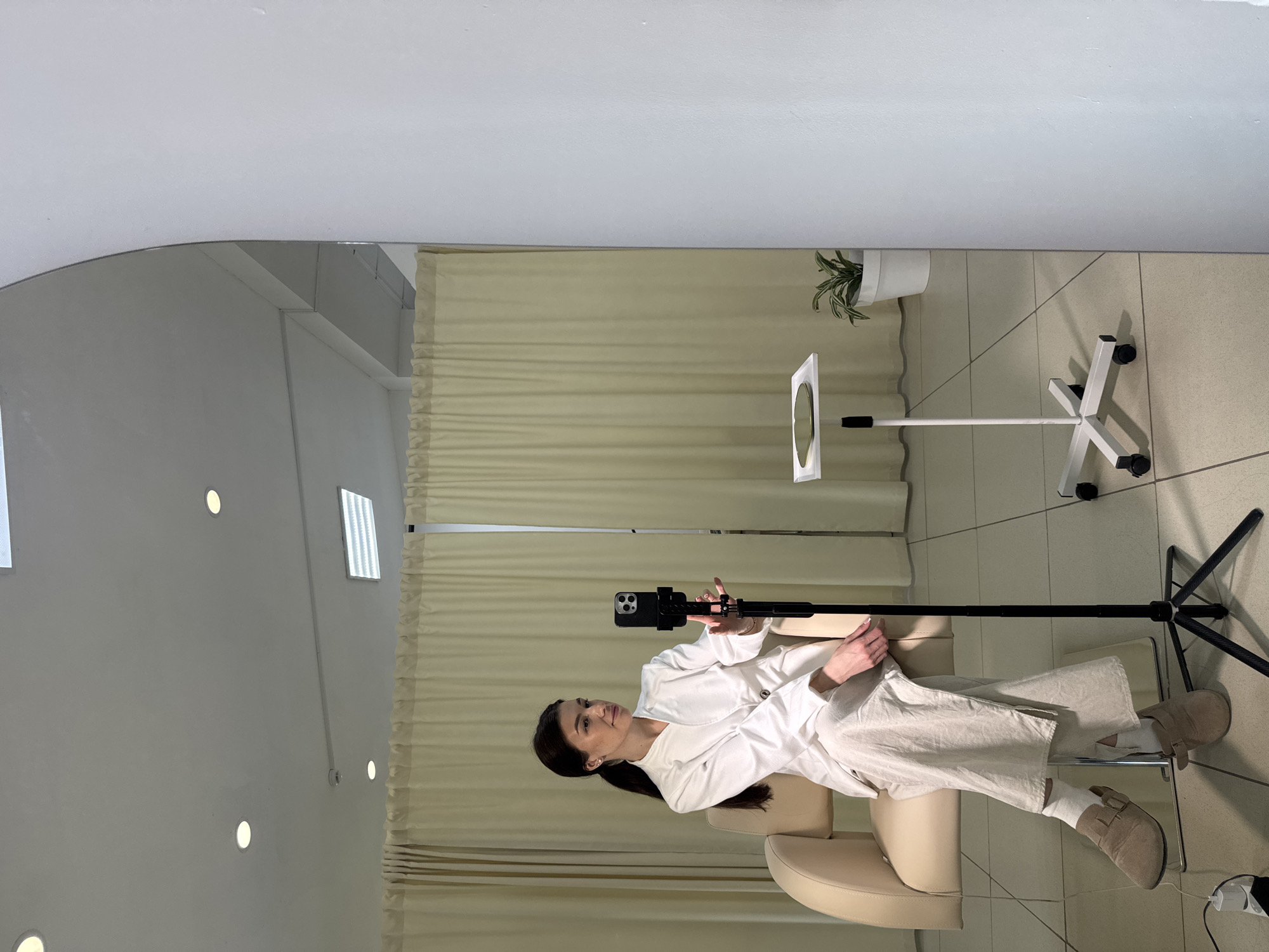 A woman with dark hair, wearing a white blazer or jacket, light beige wide-leg trousers, white socks, and light brown flat shoes, is seated in a cream-colored armchair. She is positioned towards a black smartphone, which is mounted on a black tripod directly in front of her. Her hands are near the phone, suggesting she is operating it or preparing to record.
To the woman's right, a white stand holds a circular ring light or mirror, providing illumination or visual feedback. In the background, beige pleated curtains or blinds cover a wall, and a small potted plant with green foliage is visible. The room features light-colored walls and a tiled floor. The ceiling is equipped with multiple round recessed lights and a rectangular light fixture, providing bright, even illumination.
The entire scene, situated in Chaikovsky, Russia, appears to be a reflection in a large mirror, creating a sense of depth and showcasing more of the room's interior, including additional ceiling lights. The woman maintains a composed, neutral expression, directing her gaze towards the smartphone, indicating she is likely recording a video, live-streaming, or taking a photo in a well-lit indoor studio or content creation space. No visible text is discernible in the image.