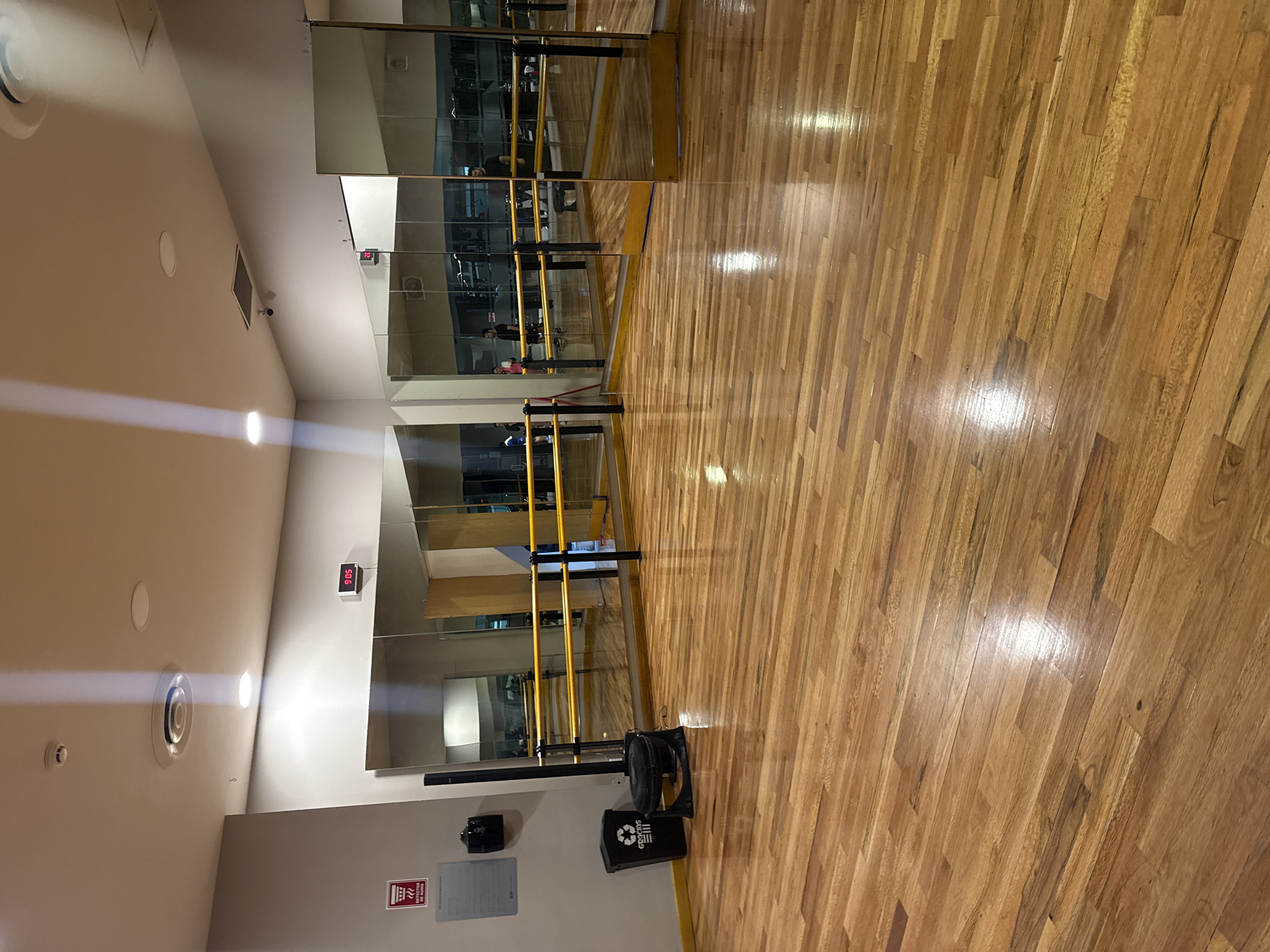 This media file captures the interior of a well-lit dance or fitness studio, likely in Puebla, Mexico, given the visible Spanish text. The dominant feature is a highly polished, light-colored wooden floor composed of rectangular planks, which reflects the overhead artificial lighting. One wall is entirely covered with large mirrors, creating reflections of the room's interior.
Along the mirrored wall, a double-railed ballet or fitness barre with yellow wooden rails and dark support posts is installed. In the reflections of the mirrors, at least three individuals in exercise or casual attire are partially visible, indicating active use of the studio.
The white ceiling features multiple recessed circular lights, circular ventilation grilles, a square vent, and a small white smoke detector or sensor. Mounted on the wall above the mirrors is a digital clock displaying "9:05" in red LED digits, and a small black security camera is also visible. Near the floor, a black industrial fan is positioned, along with a black recycling bin that clearly displays the recycling symbol and the word "GRACIAS" (Spanish for "thank you"). A red fire safety sign, featuring a fire extinguisher icon, is also affixed to the lower wall, with an illegible white sign beneath it. The setting conveys a clean, functional environment dedicated to physical activity.