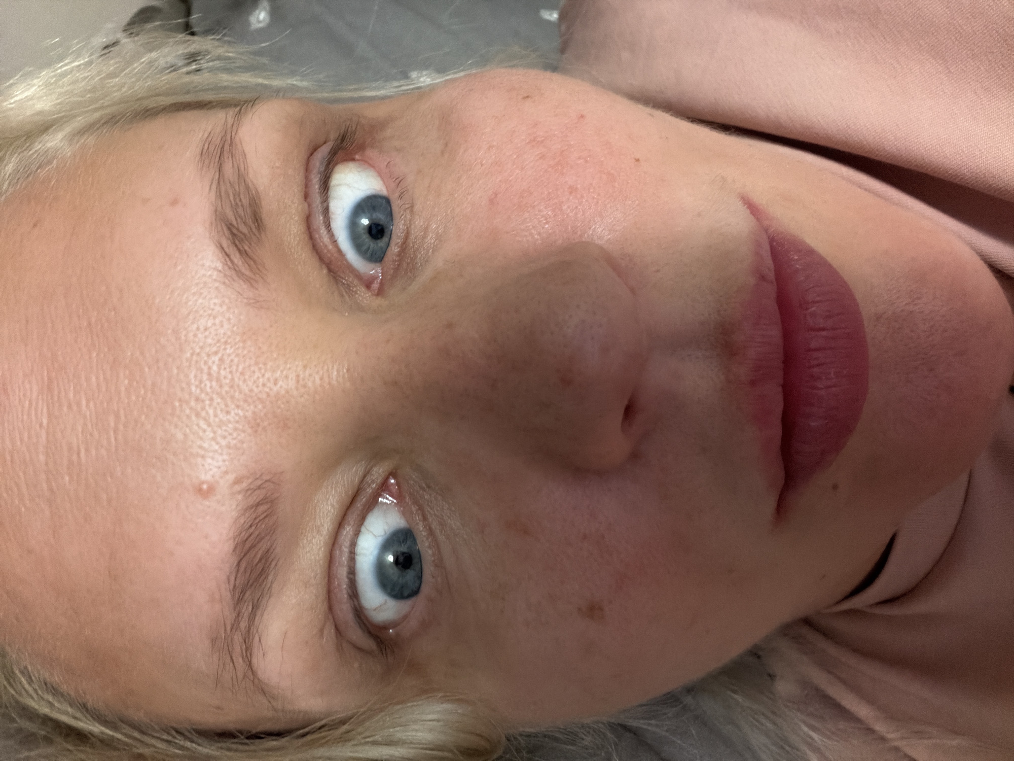 The image is a close-up, upward-angled selfie of a woman's face, taken in Kazan, Russia. The woman has light-colored hair and blue eyes, which are wide open and looking directly at the camera. Her facial expression is exaggerated, with her eyes appearing to bulge. She has a few moles and freckles visible on her skin. She is wearing a pink shirt. The background is out of focus and appears to be indoors, possibly on a bed given the texture of the surface behind her. No other people or objects are clearly discernible.