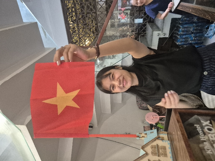 A woman holds up a Vietnamese flag, smiling at the camera. The flag is red with a yellow star. The woman wears a black shirt and a striped skirt. In the background, another person is partially visible. Nearby, a "Kha Hostel ROX" box sits on a surface along with other small objects. Stairs are visible on the left.