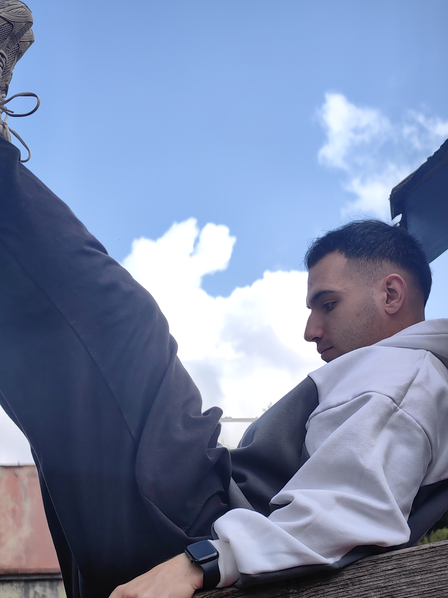 A low-angle shot of a young man in Shumen, Bulgaria. He's lying on a weathered wooden structure with his legs propped up, one foot visible against a partly cloudy sky. The man has dark hair, a light complexion, and is wearing a white and gray hoodie and dark-colored sweatpants. A smartwatch is visible on his wrist. In the background is a glimpse of a building and a rooftop structure, with hints of greenery.