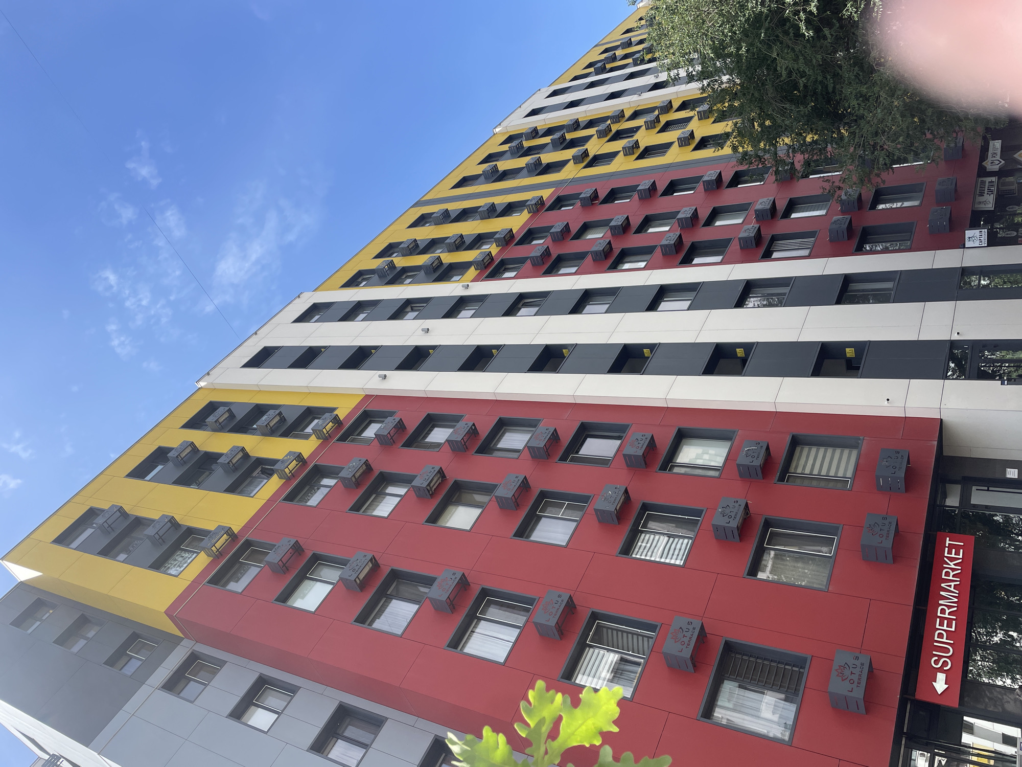 This is a low-angle, full shot of a modern, colorful apartment building on a sunny day. The building is segmented into different color blocks: gray on the left, followed by red, yellow, and then white and gray stripes towards the right. Numerous rectangular air conditioning units are mounted on the exterior walls of the red and yellow sections. Windows are visible throughout the building, some with curtains. On the ground level, a red sign with white text indicates "SUPERMARKET" with an arrow pointing left. Some greenery, likely trees and leaves, is visible in the foreground on the left and right edges of the frame, partially obscuring the lower parts of the building. The sky above is a clear blue with a few wispy clouds. The building appears to be located in the city of Almaty, Kazakhstan, as suggested by some visible Cyrillic text on signs at the ground level, such as "КАПИТАН".