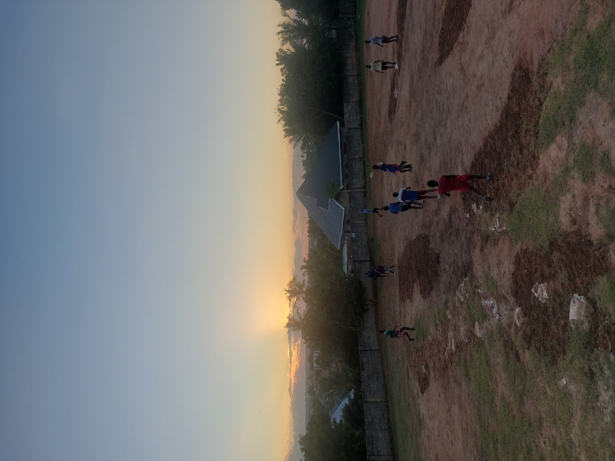 The image shows a view from a high angle, likely from a building overlooking a landscape at what appears to be sunset. In the foreground, a group of people are scattered across a partially grassy, partially dirt field; some appear to be playing sports, possibly soccer, as there are individuals in athletic wear and potentially a ball. A stone wall borders the field. Beyond the field, there are houses and trees. The sky is a gradient of blue to orange, indicating either sunrise or sunset. Based on the presence of lush vegetation and buildings, the location is likely Dar es Salaam, Tanzania, as per the prompt.