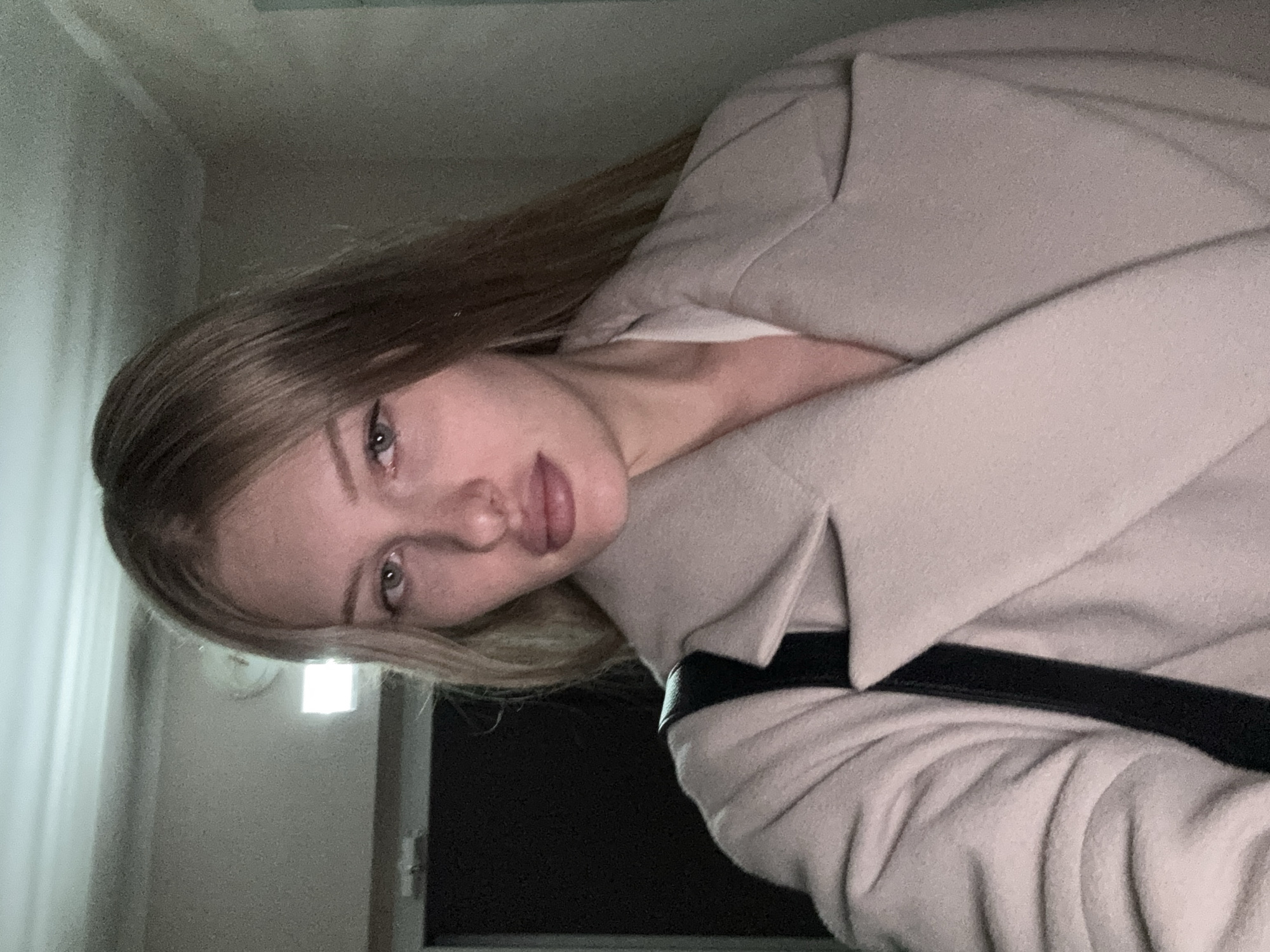 A medium shot captures a fair-skinned woman with long, blonde hair from a slightly low angle. She is looking directly at the camera, with a neutral expression. She is wearing a light-colored coat, possibly beige or cream, over a white top. A black strap, likely from a bag, is visible crossing her shoulder. The background is dimly lit and appears to be an indoor hallway. A white light fixture is visible on the ceiling, and a dark door or wall is to her left. The scene is indicative of an indoor environment, possibly a hallway in a residential building or an office space. There is no clear indication of the specific location within Kondratovo, Russia, beyond it being an indoor, man-made environment.