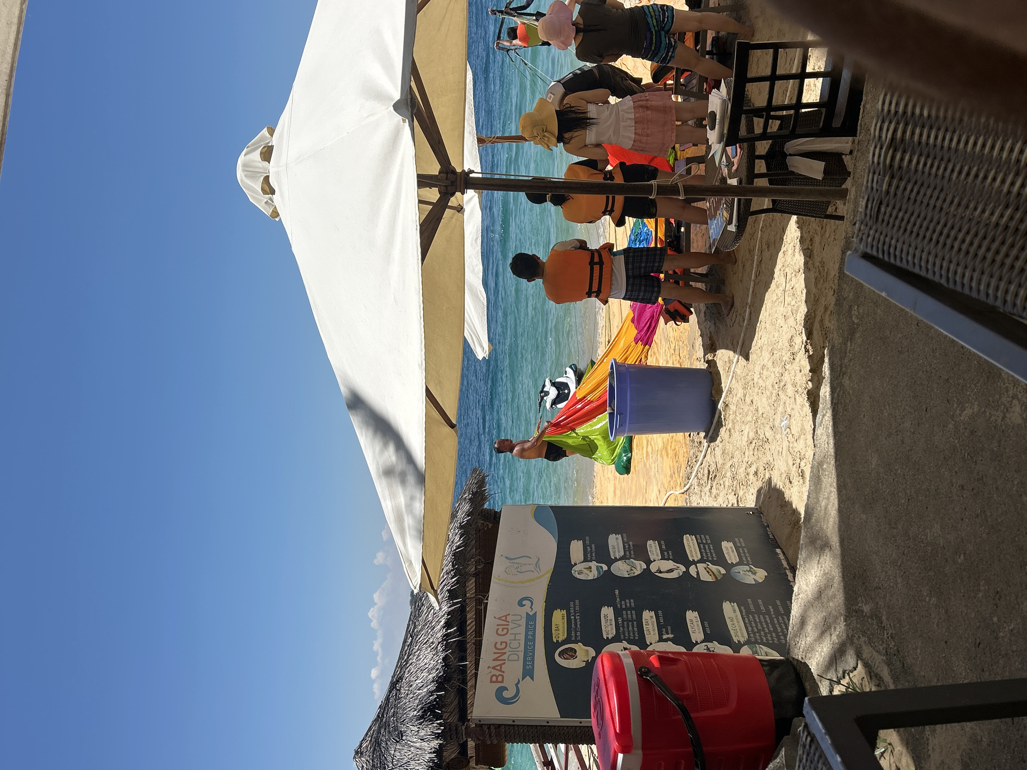 The image depicts a sunny beach scene, likely in Bac Nha Trang Ward, Vietnam, with several people engaged in recreational activities. In the foreground, a thatched-roof hut stands to the left, with a red cooler and a price list sign for services such as "DU BAY" (parasailing) and "MO TO NUOC" (jet skiing). Beyond the hut, people are visible on the sandy beach. One man in swim trunks is holding a colorful parachute or sail, and a jet ski is partially visible. Several individuals wearing orange life vests are standing near the water, possibly preparing for or observing water sports. Two people are sitting at a table under a large white umbrella, with chairs around it. The ocean is a clear blue, with gentle waves. The overall atmosphere is relaxed and suggests a tourist destination.