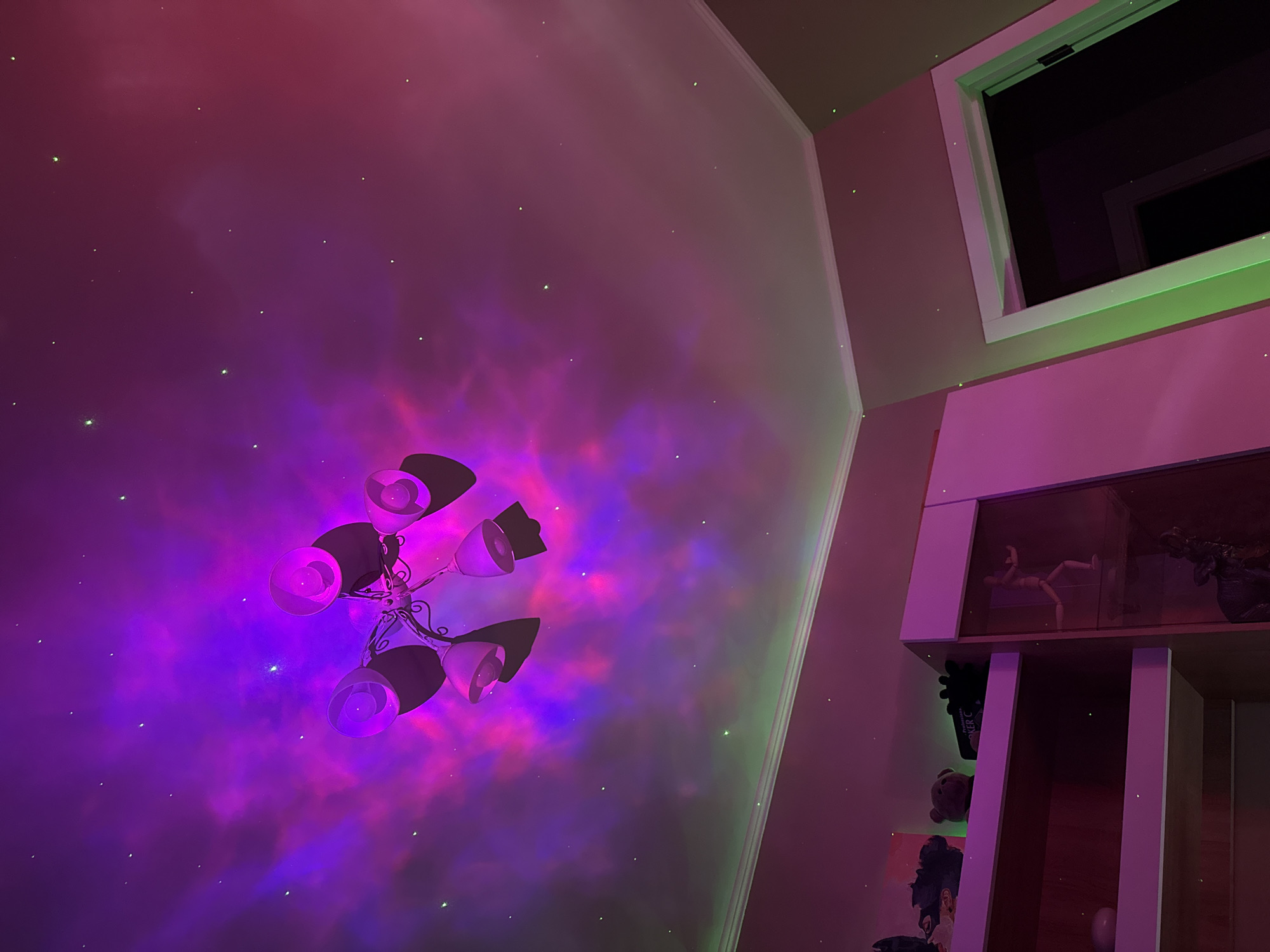 The image shows an interior space with pink and purple lighting effects. A multi-armed chandelier is positioned prominently on the wall, illuminated in bright purples and blues. The wall itself is covered with what appears to be a projected image of a nebula or galaxy-like pattern, also illuminated in shades of purple and pink, with scattered green points of light. To the right, there is a window with a white frame and a bookshelf. The bookshelf features various objects on its shelves, including what looks like a mannequin and other decorative items. The overall aesthetic is vibrant and somewhat theatrical. The specific location in the city of Unknown, Unknown is not discernible from the image.