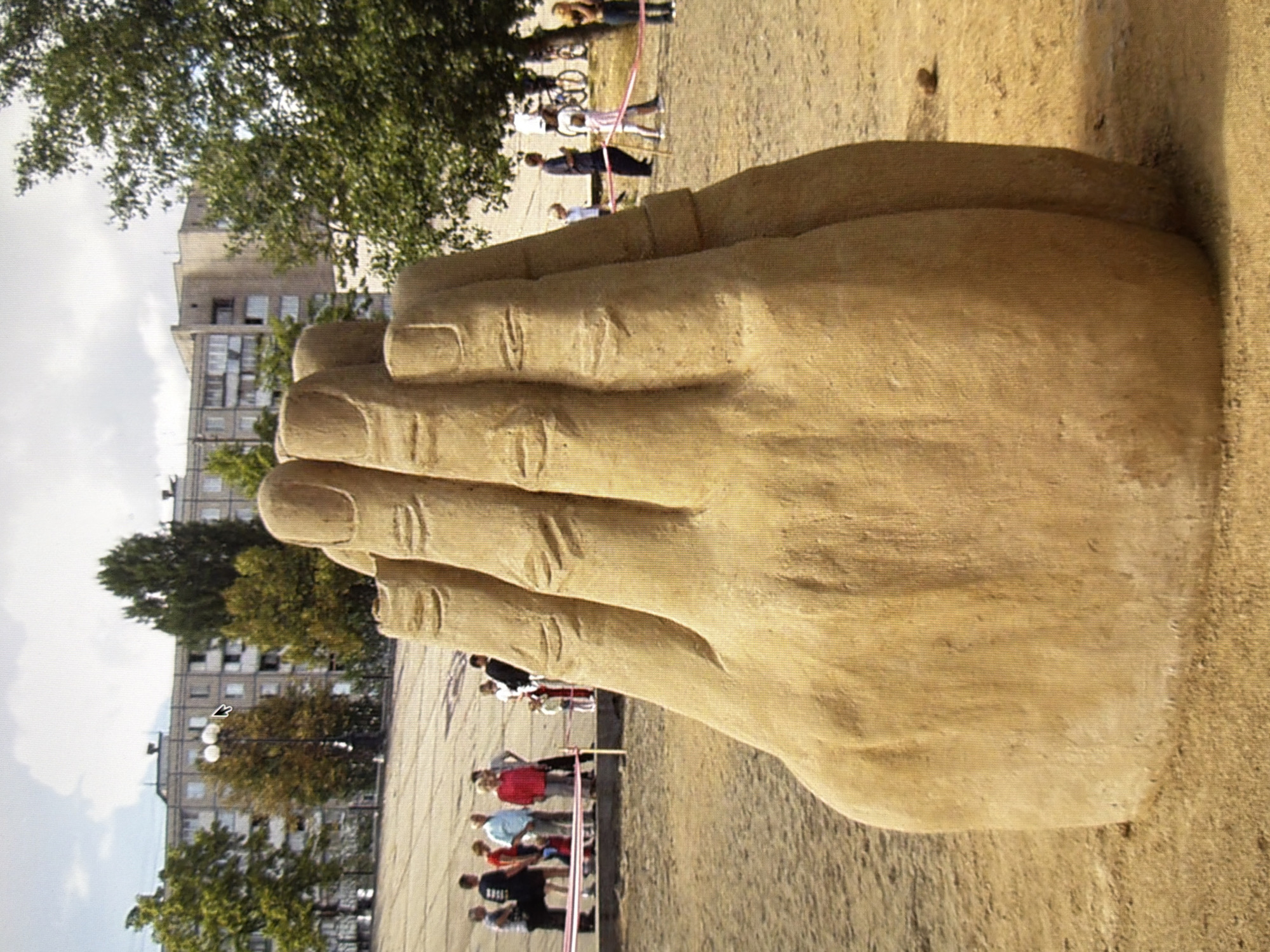 The image shows a large sculpture of a hand in what appears to be an outdoor public space. The sculpture is made of a light brown material, likely sand or concrete, and is positioned in the foreground, dominating the frame. The hand is depicted with its fingers slightly curled and is life-sized or larger than life-sized.
In the background, several people are visible, some standing and observing the sculpture, while others appear to be walking. They are dressed in casual clothing, suggesting a daytime scene. Behind the people, there are trees with green and yellow foliage, indicating an autumn season. Further in the background, there are multi-story residential buildings with a somewhat utilitarian architectural style. The sky is partly cloudy.
The overall setting appears to be a park or a public square. This sculpture is located in Kyiv, Ukraine, specifically at Independence Square (Maidan Nezalezhnosti).