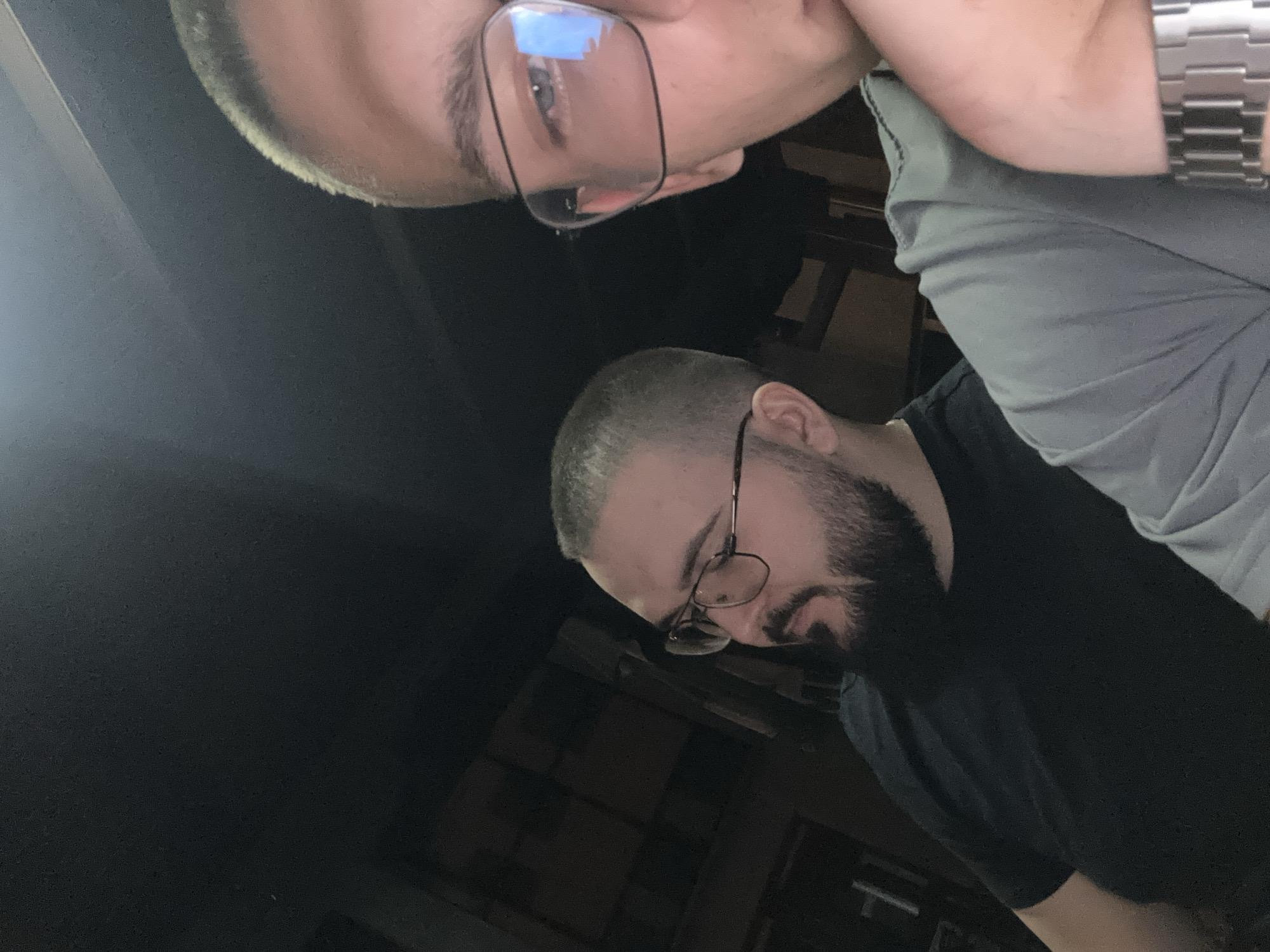 Two men are captured in a close-up, slightly elevated, dimly lit indoor photograph. The man on the left, positioned closer to the center of the frame, has short hair, a full beard, and wears glasses with thin frames. He is looking downwards and to his left. He is wearing a black t-shirt. The man on the right is partially visible, with his face in the foreground and cropped at the bottom of the frame. He is wearing glasses with thick black frames and a grey t-shirt. His gaze is directed towards the camera. A silver watch is visible on his left wrist. The background consists of dark, indistinct structures, suggesting an indoor setting with limited lighting, possibly a room or a covered outdoor area. The presence of a city name like Nedelino, Bulgaria, is not indicated by any visual elements within the image itself.