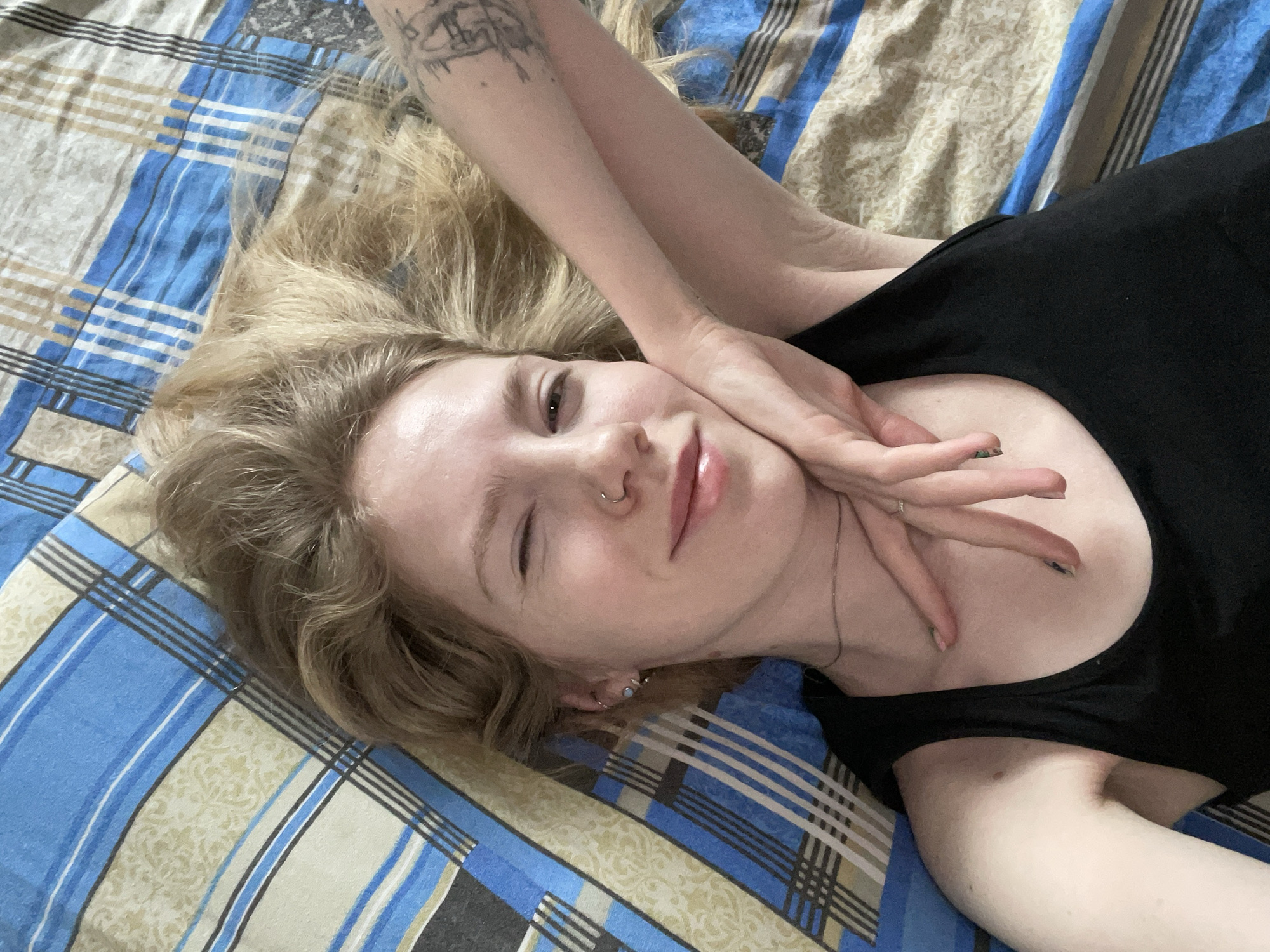 A fair-skinned individual with blonde, wavy hair is lying supine on a bed with patterned bedding, presenting a selfie-style portrait. The person is smiling, with their left eye closed in a wink and their right eye open. Their left hand, adorned with black nail polish on some fingers and a ring on the ring finger, rests near their chin. The individual wears a black tank top and a thin chain necklace. Visible piercings include a silver nose hoop and multiple left ear piercings, including a light-colored stud. A black ink tattoo with abstract elements is present on the upper left arm. The bedding features a plaid pattern of blue, beige, black, and white stripes. The scene is located within a private residence in the city of Izhevsk, Russia.