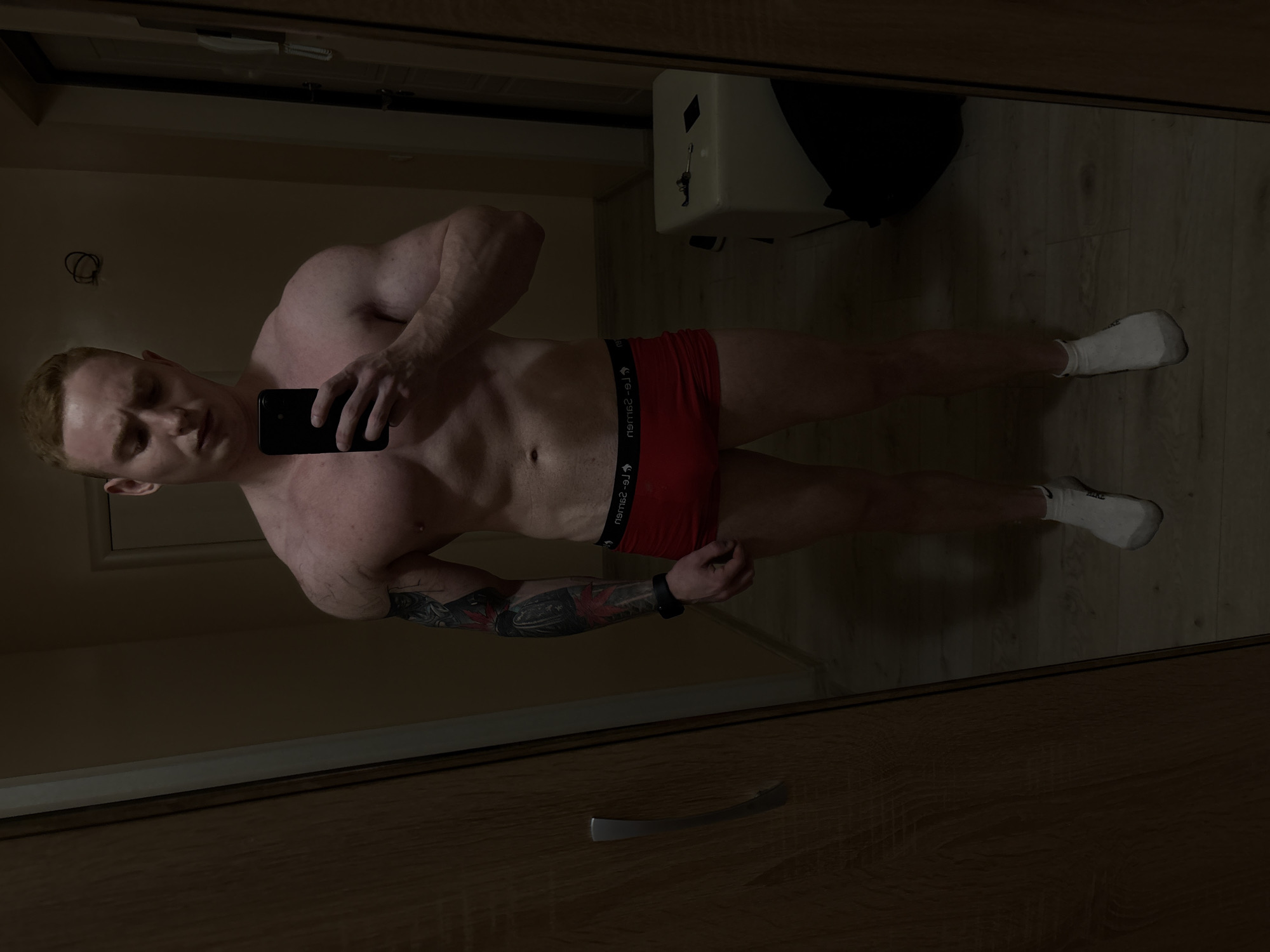 A mirror selfie, taken indoors, features a muscular, light-skinned man with reddish-blonde hair. He stands wearing red boxer briefs and white socks, with a dark smartwatch on his left wrist. His left arm is adorned with a detailed tattoo sleeve. The man holds a dark-colored smartphone in his right hand, positioned to capture the reflection.
The background, seen in the mirror, shows a wooden floor and a dark wooden cabinet or wardrobe with a silver handle. A light-colored appliance, possibly a water heater, is visible on the upper right of the reflection, with some dark material atop it. The dim lighting suggests an evening or nighttime setting within a home, consistent with an interior in Chaikovsky, Russia. The waistband of his boxer briefs displays a repeated, partially mirrored brand name, appearing as "r9-2a9m2u".