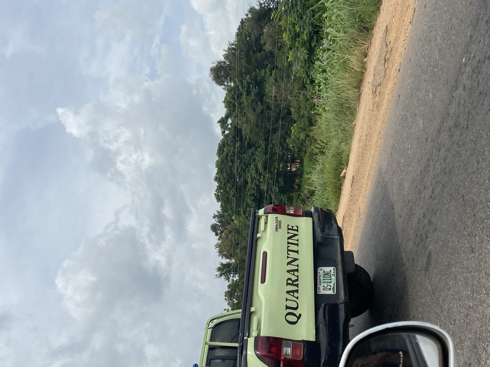 A light green pickup truck with "QUARANTINE" written in black on its tailgate is parked on the side of a road in Riyom, Nigeria. The truck has a license plate that reads "QS-110NC". The truck is surrounded by lush green vegetation, including trees and what appears to be a cornfield. The sky is overcast with scattered clouds. In the foreground, a portion of a side mirror is visible, reflecting a person. The road is paved and shows signs of wear and tear.