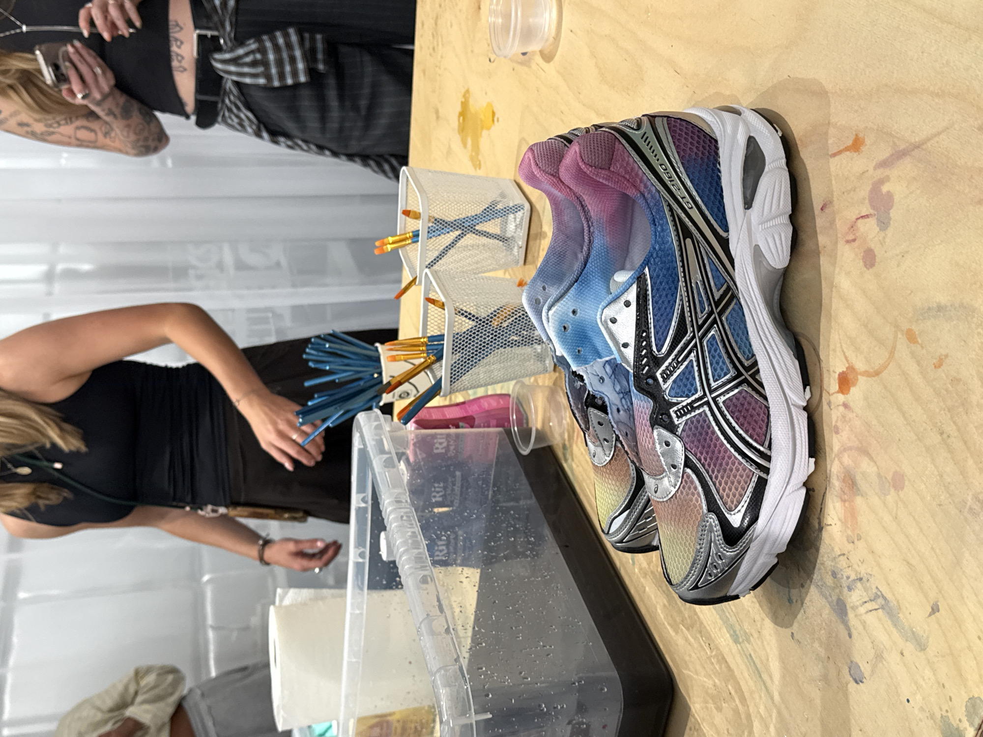 The image shows a craft table with a pair of colorfully dyed Asics GT-2160 running shoes, a container of dark liquid, paintbrushes, and other craft supplies. Two women are visible, one with a black tank top and another wearing a black tank top with a striped garment around her waist. The location is potentially an art studio or crafting space.