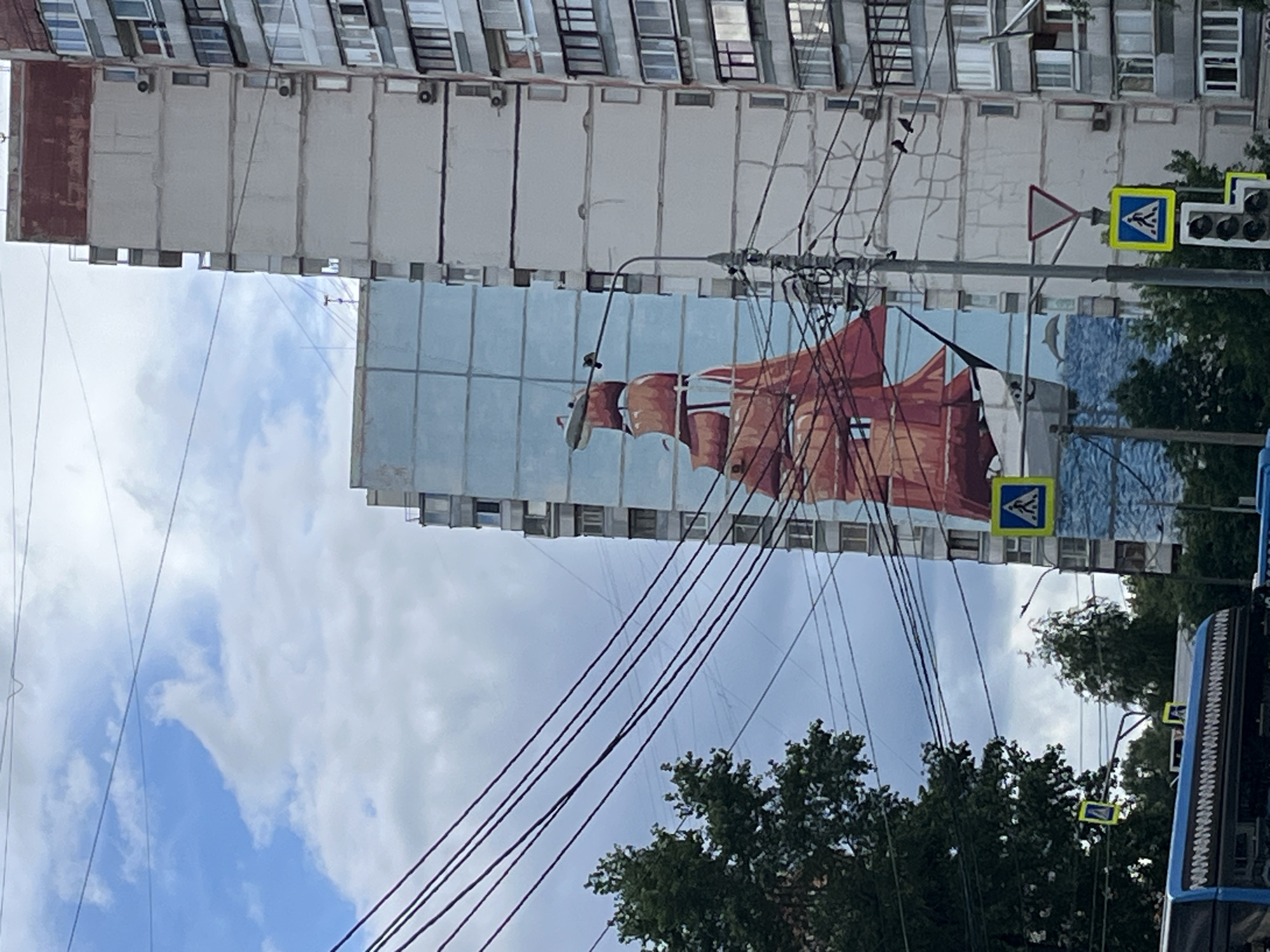 An outdoor, daytime, medium shot captures the side of a residential building in Moscow, Russia, adorned with a large mural of a tall ship with orange sails. The mural, painted on a section of the building's exterior, depicts the ship sailing on a blue sea, with a dolphin leaping from the water. Overhead, a network of thick, dark wires stretches across the frame, intersecting the building and the mural. Adjacent to the mural, another building is visible, its facade a light gray with numerous windows. Several traffic signs are present: a pedestrian crossing sign with a blue background and a white walking figure, and a triangular red-bordered sign. Trees with green foliage are seen in the lower left and right portions of the image, and a portion of a blue bus is visible at the bottom. The sky is a mix of blue with scattered white clouds.