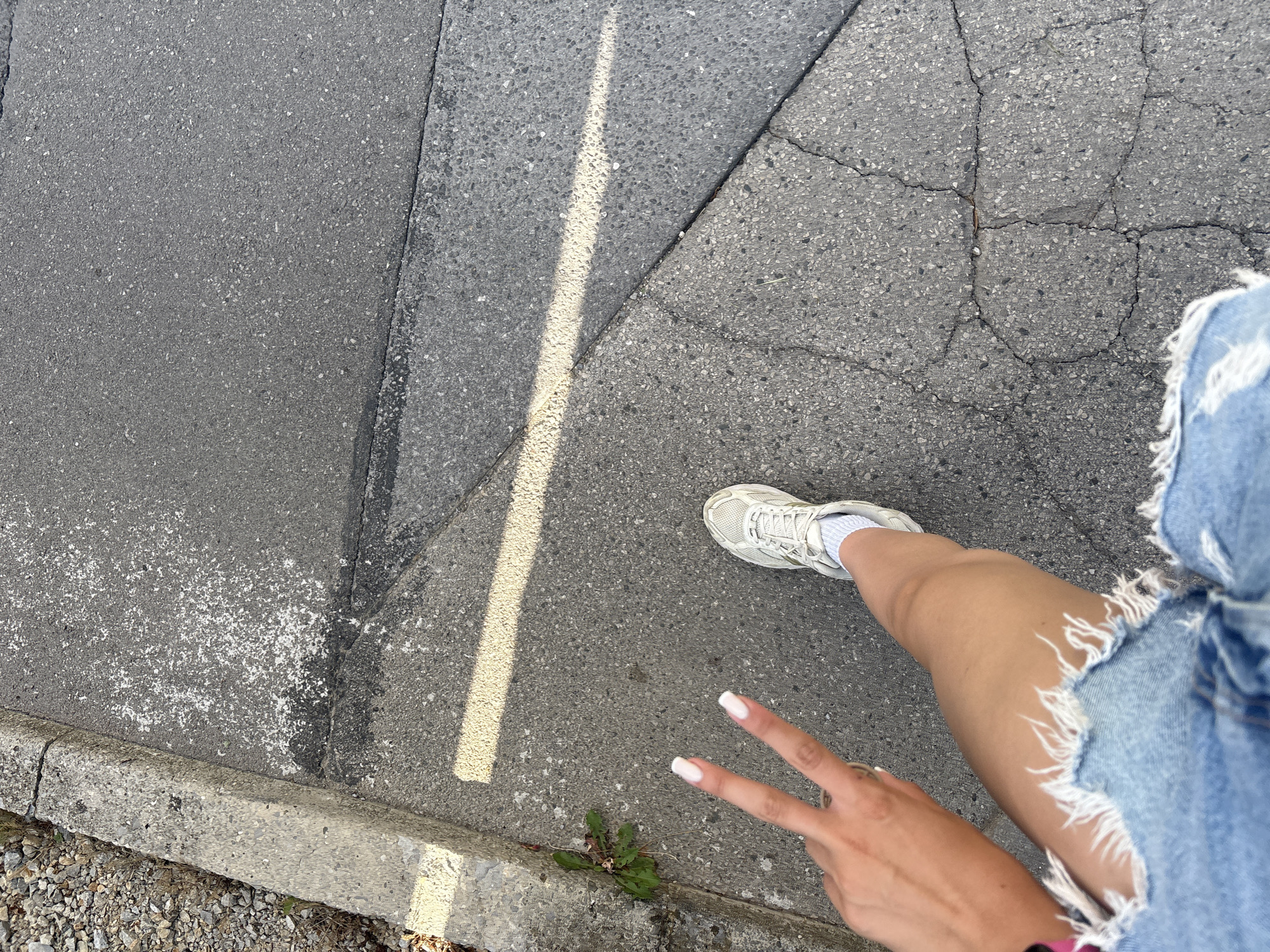 The image shows a person's leg and hand, as they are walking on a cracked asphalt sidewalk. The person is wearing denim shorts and a white sneaker with white socks. The hand is making a peace sign, and has white nail polish on. Sunlight is casting a shadow across the sidewalk. The sidewalk is next to a concrete curb, and a patch of gravel.