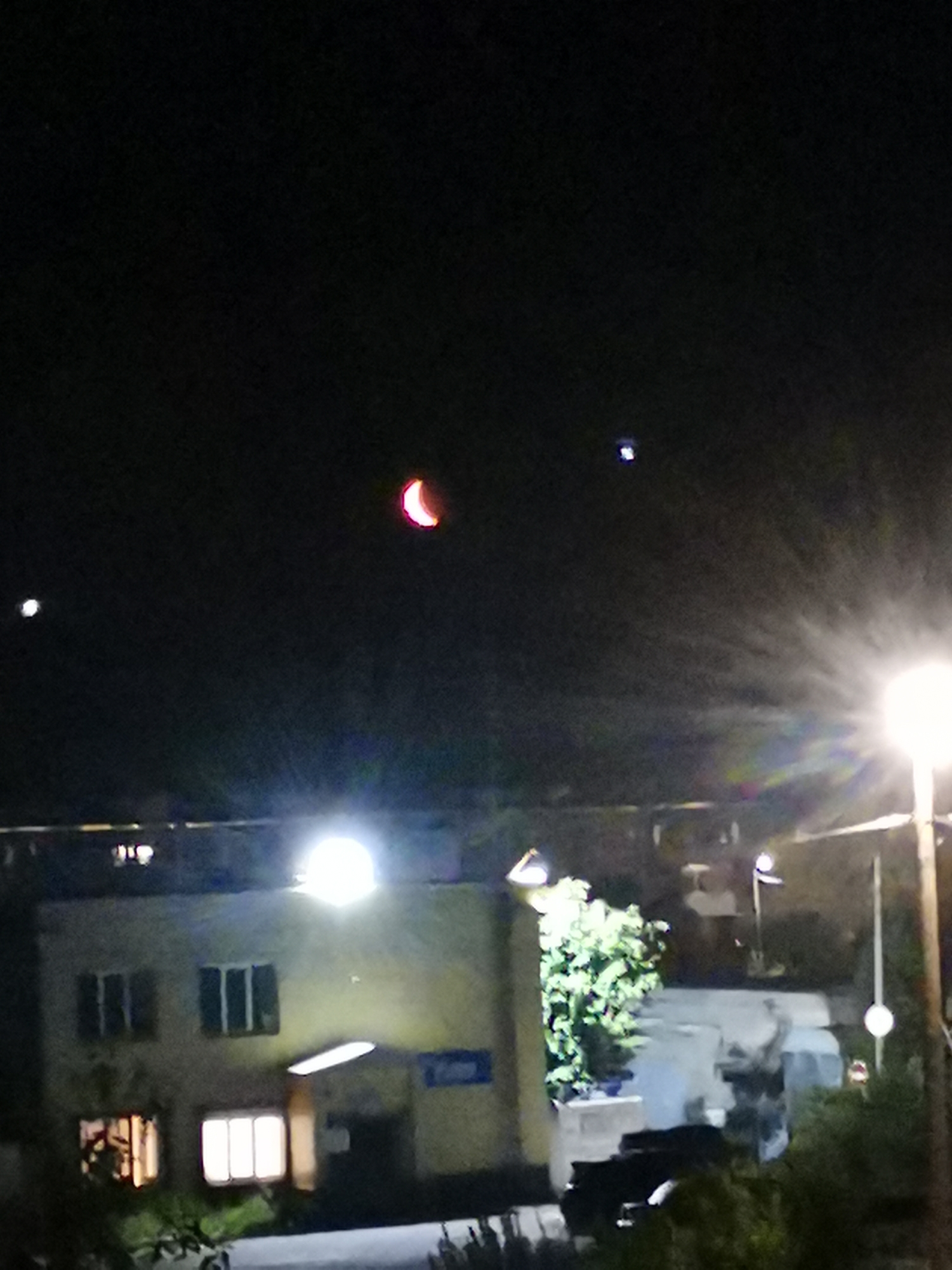 The image captures a night scene in Kizel, Russia, featuring a crescent-shaped celestial body with a reddish-orange hue, possibly the moon during an eclipse or an atmospheric phenomenon, positioned high in the dark sky. To the left of this celestial body, a smaller, bright white point of light is visible. Further to the left and lower down, another white point of light appears near the horizon.
In the foreground, a low-rise building with illuminated windows is visible. The building's facade is a light color, and the windows emit a warm, yellowish light from within. A prominent, bright light source, possibly a street lamp, shines brightly from the right side of the frame, casting rays of light that create a starburst effect and illuminate the surrounding area. The base of a pole supporting this light source is also visible.
The lower portion of the image shows the silhouettes of trees and what appears to be a dark vehicle parked or driving on a road. The overall ambiance is dark, typical of a nighttime setting, with artificial lights from the building and street lamp providing the primary illumination for the lower part of the scene. The focus appears to be on the celestial event and the illuminated elements of the urban landscape.