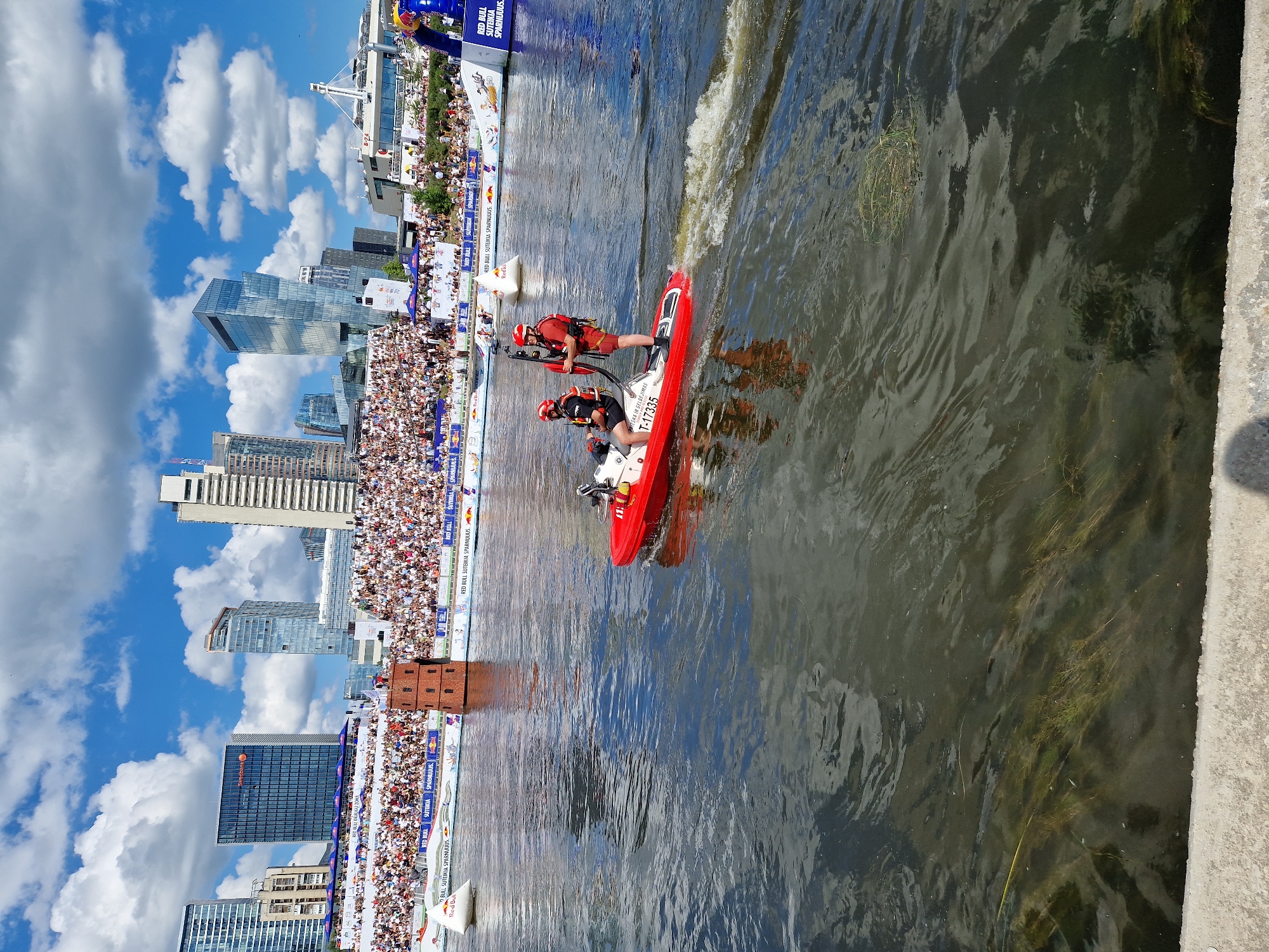 The image depicts a scene on the Neris River in Vilnius, Lithuania, during what appears to be a public event. In the foreground, a red and white jet ski is moving through the water, leaving a wake. Two individuals wearing life vests and helmets are on the jet ski; one is operating it, and the other is standing and holding a pole. In the background, a large crowd of spectators is seated in tiered stands along the riverbank. Behind the stands, the Vilnius skyline is visible, featuring modern skyscrapers and other buildings. A notable landmark in the middle ground is a small, brown, brick tower structure situated in the water. The sky is blue with white, puffy clouds. The event seems to be branded with "Red Bull" logos visible on signage and floating buoys.
