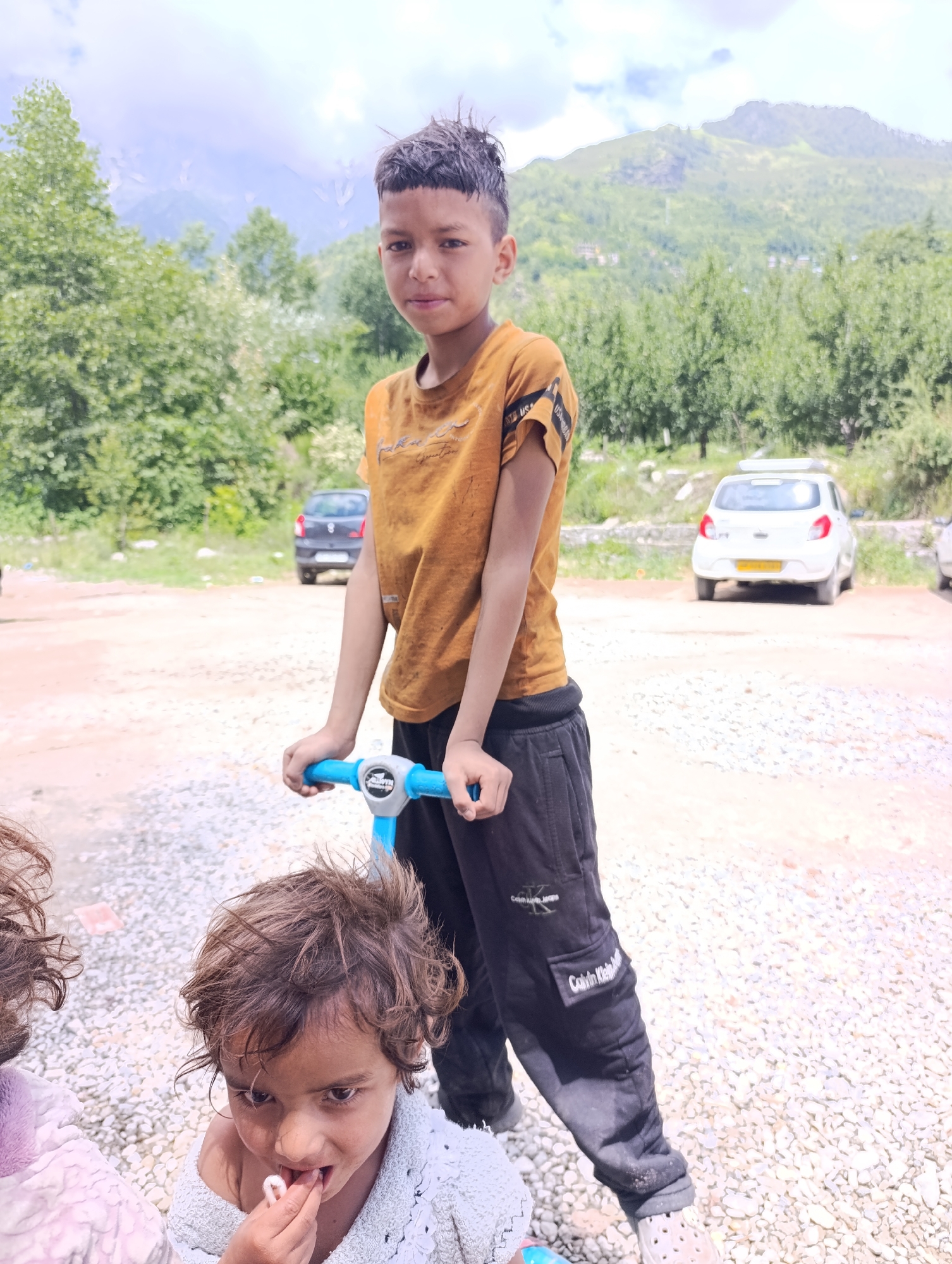 In Shanag, India, a young boy, approximately 10-12 years old, stands holding the handlebars of a blue toy scooter. He wears a mustard yellow t-shirt with a graphic print and black pants with a Calvin Klein logo. In the foreground, a younger child, with curly brown hair, is partially visible, chewing on a white object, and wearing a light blue garment. The background features a dirt and gravel-covered parking area, with trees and mountains in the distance under a cloudy sky. Two cars are parked in the background, a dark colored one on the left and a white one on the right.