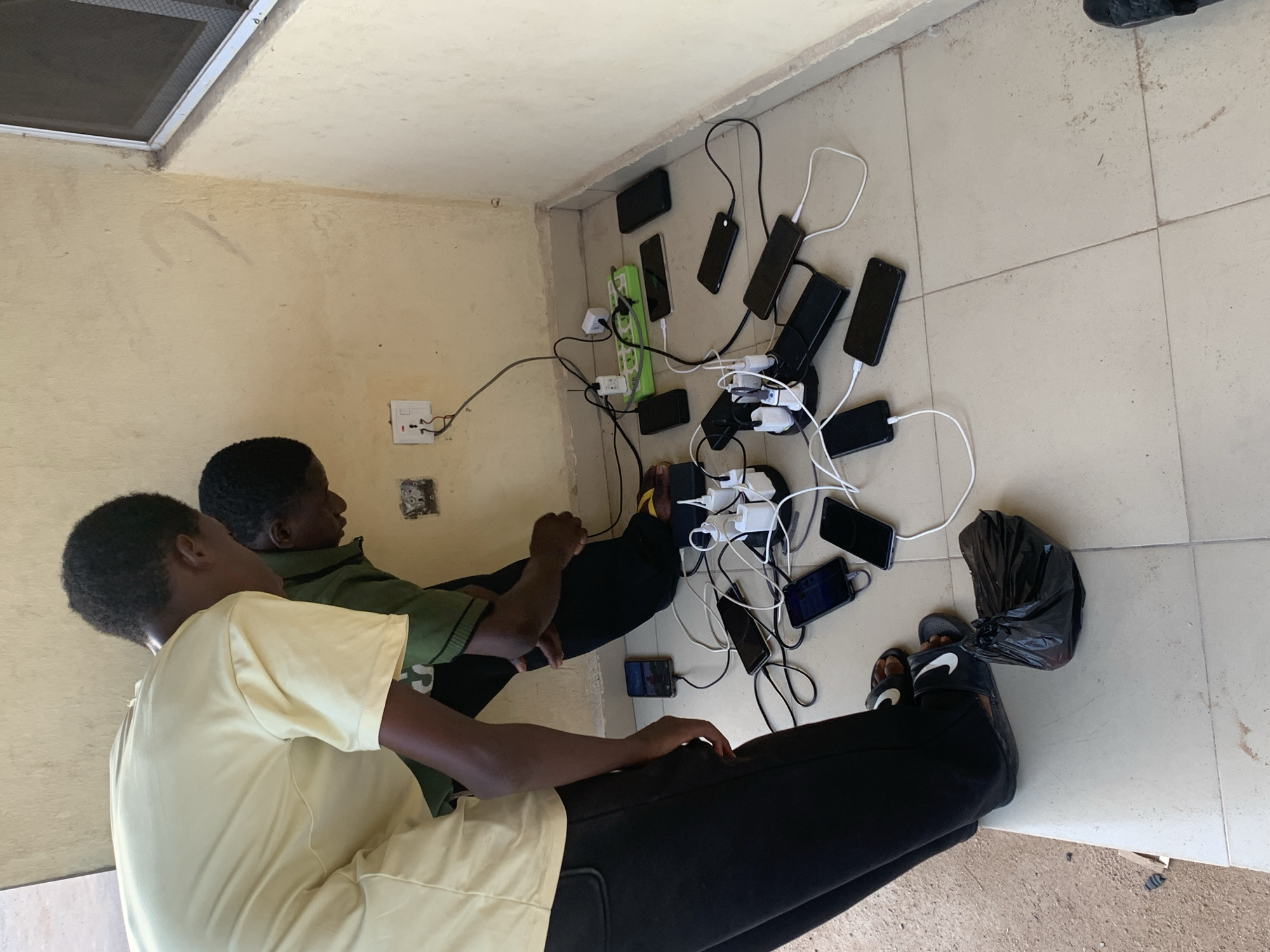 Two young men are seated on a light-colored tiled floor next to a wall in an indoor setting, likely in Lokoja, Nigeria. The man in the foreground wears a light yellow t-shirt and black trousers, while the man behind him wears a green striped polo shirt and black trousers. Both appear to be observing or managing a collection of numerous mobile phones and charging equipment.
The primary activity depicted is the charging of multiple smartphones. A single wall outlet is visible on the light beige wall, from which an extension cord connects to at least two power strips—one long green one and another black one. Many smartphones, possibly over ten, are laid on the floor, each connected via a charging cable and adapter to these power strips. The phones are mostly dark-colored and appear to be in active charging mode, indicated by the connected cables and adapters. The scene highlights a communal or shared charging arrangement, potentially due to limited access to power outlets.
Notable details include the somewhat worn appearance of the light-colored walls and ceiling, with some staining visible on both. A window with a mesh screen is visible in the upper left corner of the room. One of the men wears black Nike slides, with the distinctive white "Nike" text and swoosh logo clearly visible on the strap of one slide. A black plastic bag is positioned near his feet. The overall lighting suggests it is daytime. No specific emotions are prominently displayed by the individuals, who appear focused on the charging process.