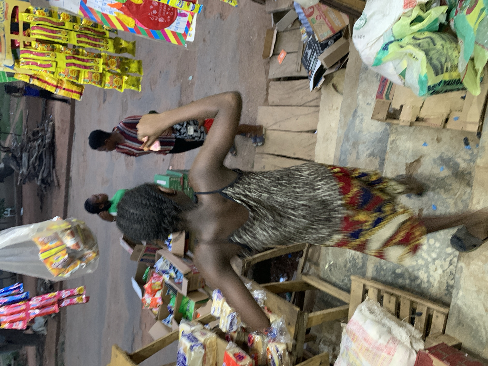 The image captures an outdoor market stall or small shop, likely in Kabba, Nigeria, as indicated by the context. The scene is bustling with various goods displayed on makeshift wooden shelves and pallets.
In the foreground, a dark-skinned woman with dark, possibly braided or natural hair, is visible from the waist up. She wears a patterned dress, with a plain dark upper section and a vibrant lower skirt adorned with red, yellow, blue, and white geometric or floral motifs. Her left arm is raised, possibly in motion or reaching for something. She wears dark, simple footwear.
To the left of her, another person, also dark-skinned, is partially visible, wearing a red, white, and dark-striped shirt. They appear to be looking at or interacting with merchandise. Further left, a display rack holds numerous yellow packaged snacks, clearly identifiable as "INDOMIE" instant noodles. Behind these, a colorful poster or calendar with strips of red, yellow, blue, and green is visible, showing numbers like "24" and "25," and partial text such as "Blue B..." and "Shuga...". A pile of wood or branches lies on the far left ground.
To the right of the main woman, and extending into the background, the area is filled with stacks of cardboard boxes, white and green plastic bags, and numerous packaged items, including loaves of bread in clear plastic wrappers. These are arranged on wooden pallets and crates, forming display stands. Another person in a green t-shirt is partially seen in the background, amidst more goods.
The ground is a rough, uneven surface, appearing to be dirt or worn concrete. The lighting suggests it is daytime, possibly overcast or within a shaded area, as there are no harsh shadows. The overall impression is one of a busy, local trading environment.