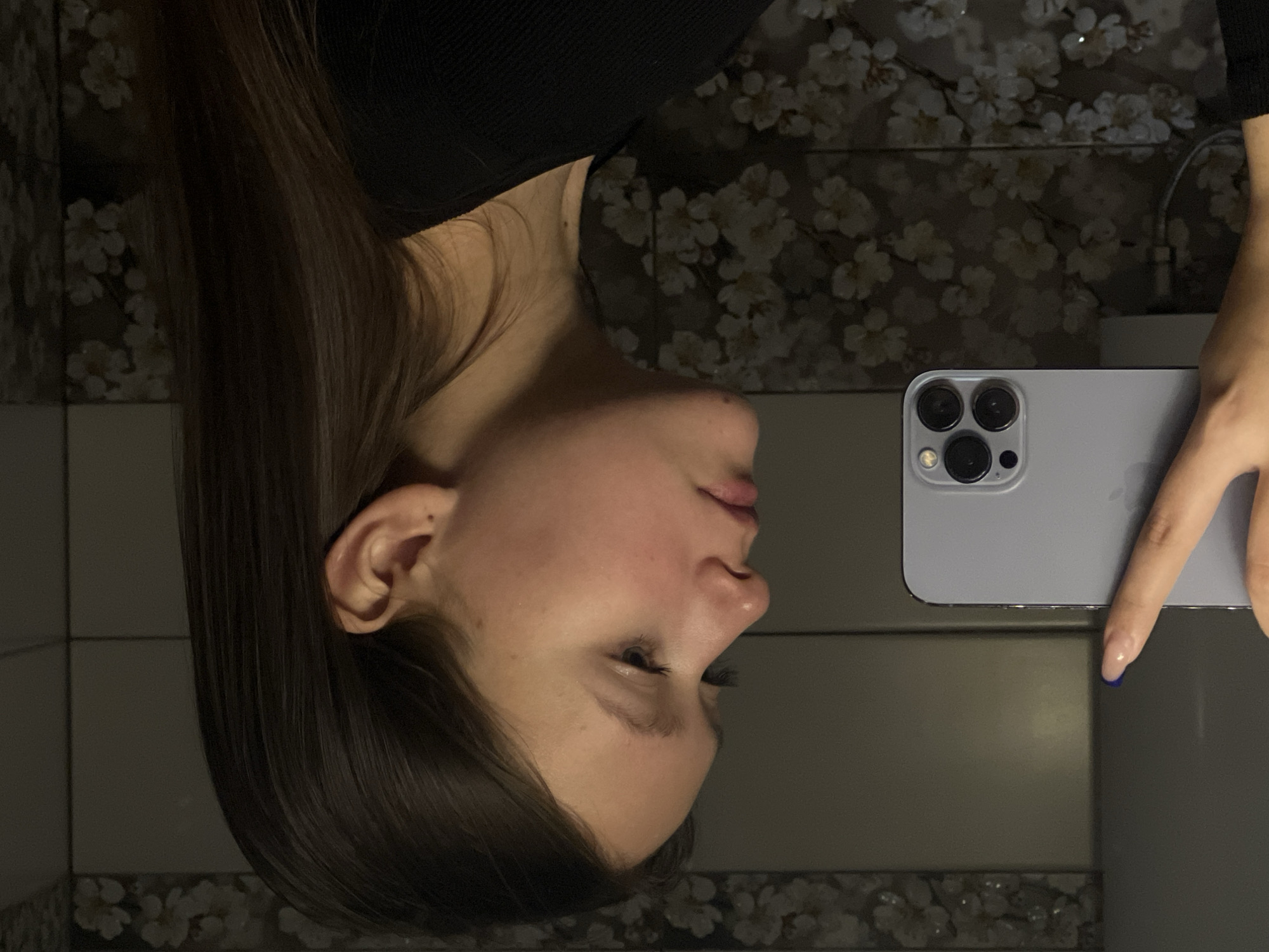 This image captures a mirror selfie of a young woman in an indoor setting, identified as Chaikovsky, Russia. The woman is seen in profile, her head slightly tilted downwards, revealing her long, straight dark brown hair draped over her shoulder. She is wearing a dark, possibly black, ribbed top. Her skin appears clear, with gentle highlights and shadows suggesting artificial or dim lighting.
She holds a light-colored smartphone, consistent with an iPhone 13 Pro or 14 Pro, in her right hand. The phone is identifiable by its distinct triple-camera module and flash. Her manicured fingers are visible gripping the device, with one fingernail (on her ring finger) adorned with dark blue polish, while others feature a natural or light nude polish.
The background reflected in the mirror is a dark wall or wallpaper, decorated with a repeating pattern of delicate white or cream floral designs, possibly cherry blossoms. To the left, plain, light-colored square tiles are visible, suggesting the setting is likely a bathroom. The overall lighting is subdued, creating a calm and intimate atmosphere. The woman's expression is neutral and focused on her reflection or the phone screen. There is no visible text within the image.