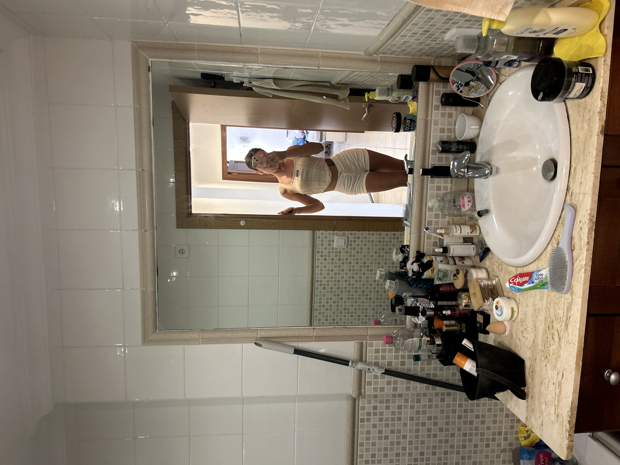 A woman is taking a mirror selfie in a bathroom located in Cala Lliteres, Spain. The bathroom features tiled walls, a vanity with a sink, and a large mirror. The woman is wearing a light-colored crop top and shorts. On the vanity, there are various toiletries, including bottles, brushes, and a tube of toothpaste. A mop is leaning against the wall to the left of the vanity. The background of the mirror shows an open doorway revealing another room with a bed.