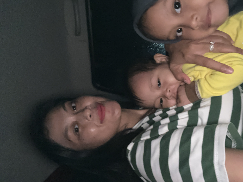 A medium close-up photograph depicts three individuals inside a vehicle at night in Tambakromo, Indonesia. An adult female with dark hair and light skin, wearing a green and white horizontal striped short-sleeved top and red lipstick, occupies the left-to-center foreground. A ring with a clear stone is visible on her left ring finger. She is holding an infant, dressed in a yellow long-sleeved top, who has one hand positioned near its mouth. To the infant's right, a young child wearing a dark blue or gray hijab is partially visible. All three subjects are looking directly at the camera. The background is uniformly dark, consistent with an enclosed vehicle interior in low light conditions.