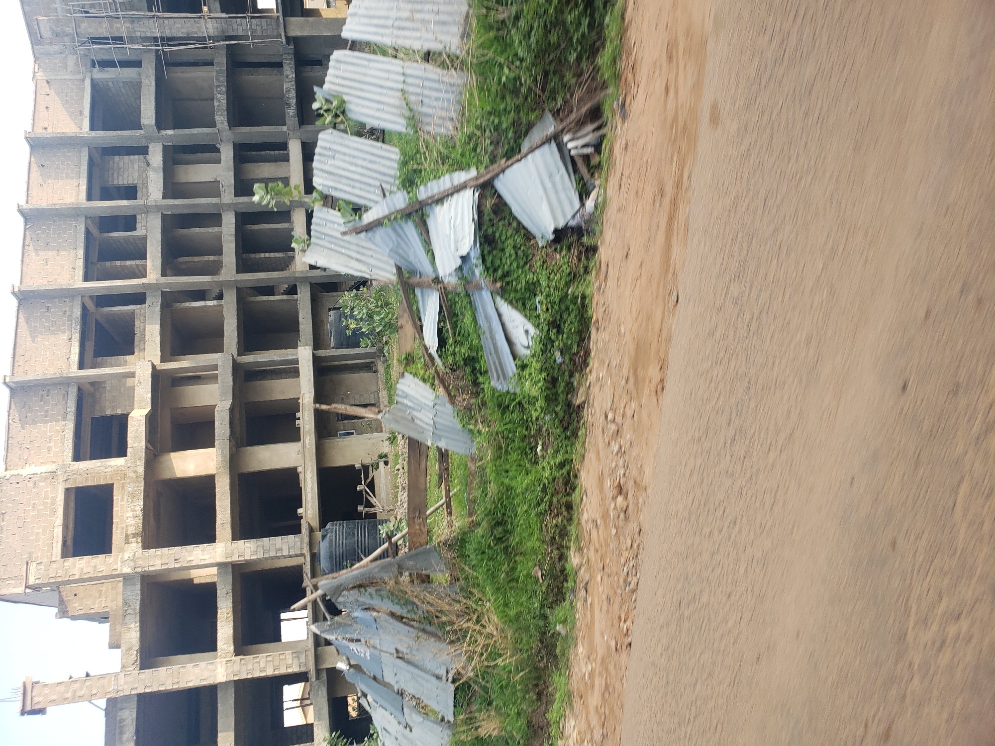 The image displays an outdoor scene featuring an unfinished multi-story building, likely a construction site. The building is a concrete structure with visible pillars, beams, and multiple floor levels, indicating it is still in the framing stage. Many window and door openings are empty, and some sections show walls made of concrete blocks.
In front of the building, a barrier constructed from corrugated metal sheets (zinc) appears to be in disarray, with several sheets scattered, bent, or partially collapsed amidst green grass and sparse bushes. A black plastic water tank is partially visible near the base of the building, behind the metallic sheets.
To the right, an unpaved road, composed of dirt and some gravel, runs alongside the construction area. The lighting suggests it is daytime, with bright, clear conditions. No people or active construction work are visible, creating a sense of a pause or halt in development. The overall setting implies a developing area, consistent with the provided location context of Narayi, Nigeria. There is no visible text in the image.