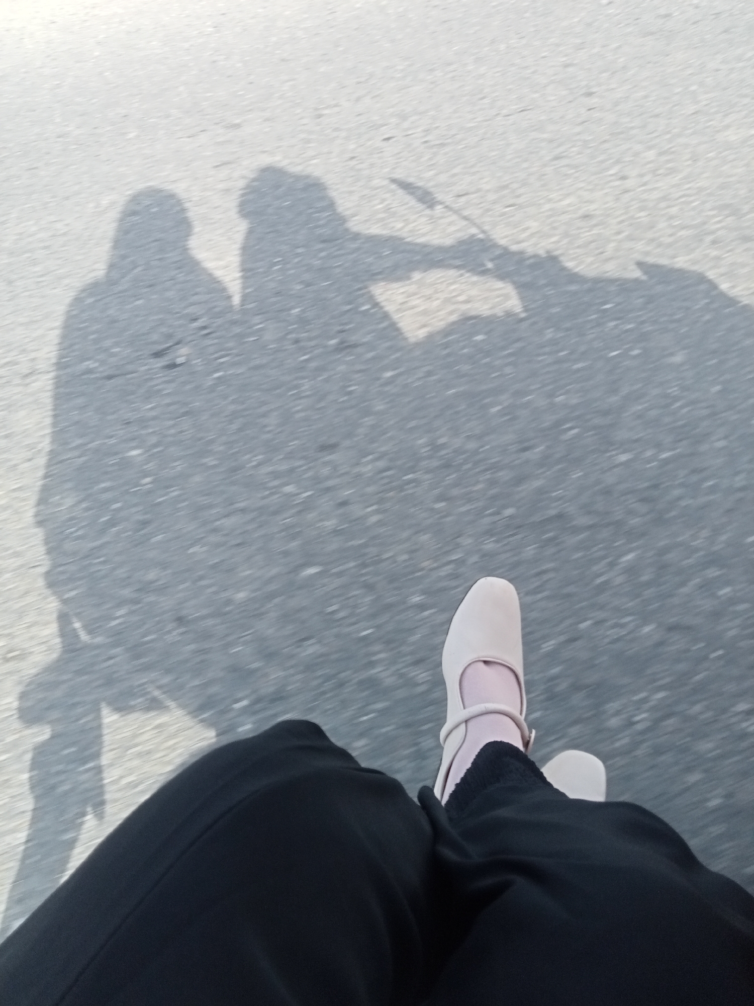 The image shows a low-angle view of two shadows cast on a paved surface, likely a road. One shadow on the left appears to be of a person wearing a headscarf, and another shadow to its right is partially obscured. In the foreground, the legs and feet of a person wearing black pants and light-colored Mary Jane shoes are visible. The shoes have a strap around the ankle. The scene suggests movement, possibly on a motorcycle or bicycle, given the elongated nature of the shadows and the blurry background. The paved surface shows a texture of small stones and asphalt. The location is identified as Matangpanyang, Indonesia.