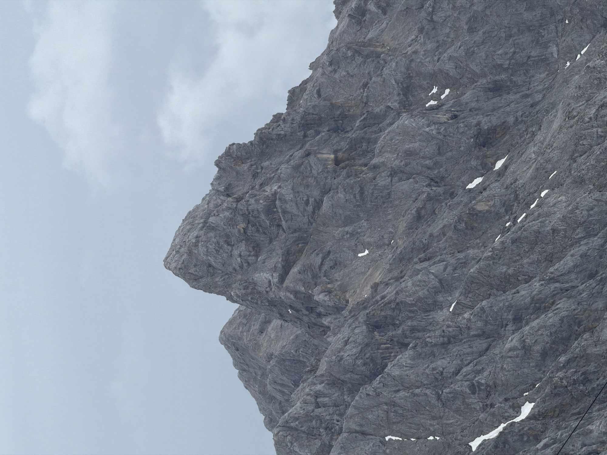 The image shows a close-up view of the jagged, rocky slopes of the Zugspitze mountain range. The mountain is predominantly grey with visible layers and textures in the rock face. Patches of snow are scattered across the slopes, particularly in shaded crevices and depressions. The sky above is overcast with soft, grey clouds, creating a diffused light that casts no harsh shadows. The overall impression is one of a rugged, natural alpine landscape under a cloudy sky. The Zugspitze is located in the Wetterstein Mountains, on the border between Germany and Austria, and is a significant landmark near Garmisch-Partenkirchen, Germany.