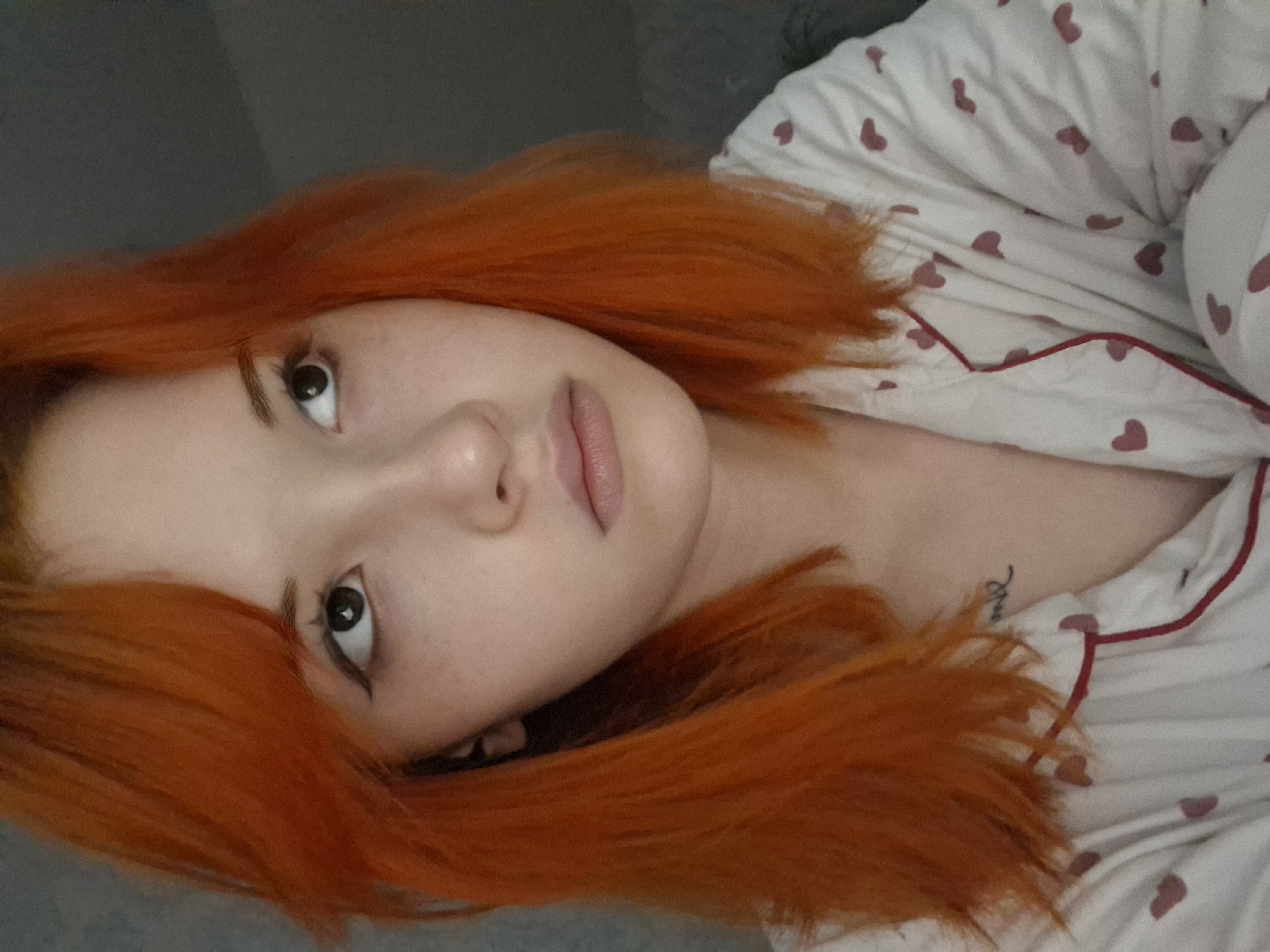 A young woman with orange hair is lying down, possibly on a bed or couch, wearing white pajamas with small red hearts. She is facing the camera, looking slightly upwards. The environment appears to be indoors, possibly a bedroom. The image does not provide any indication of the location being Chaikovsky, Russia.