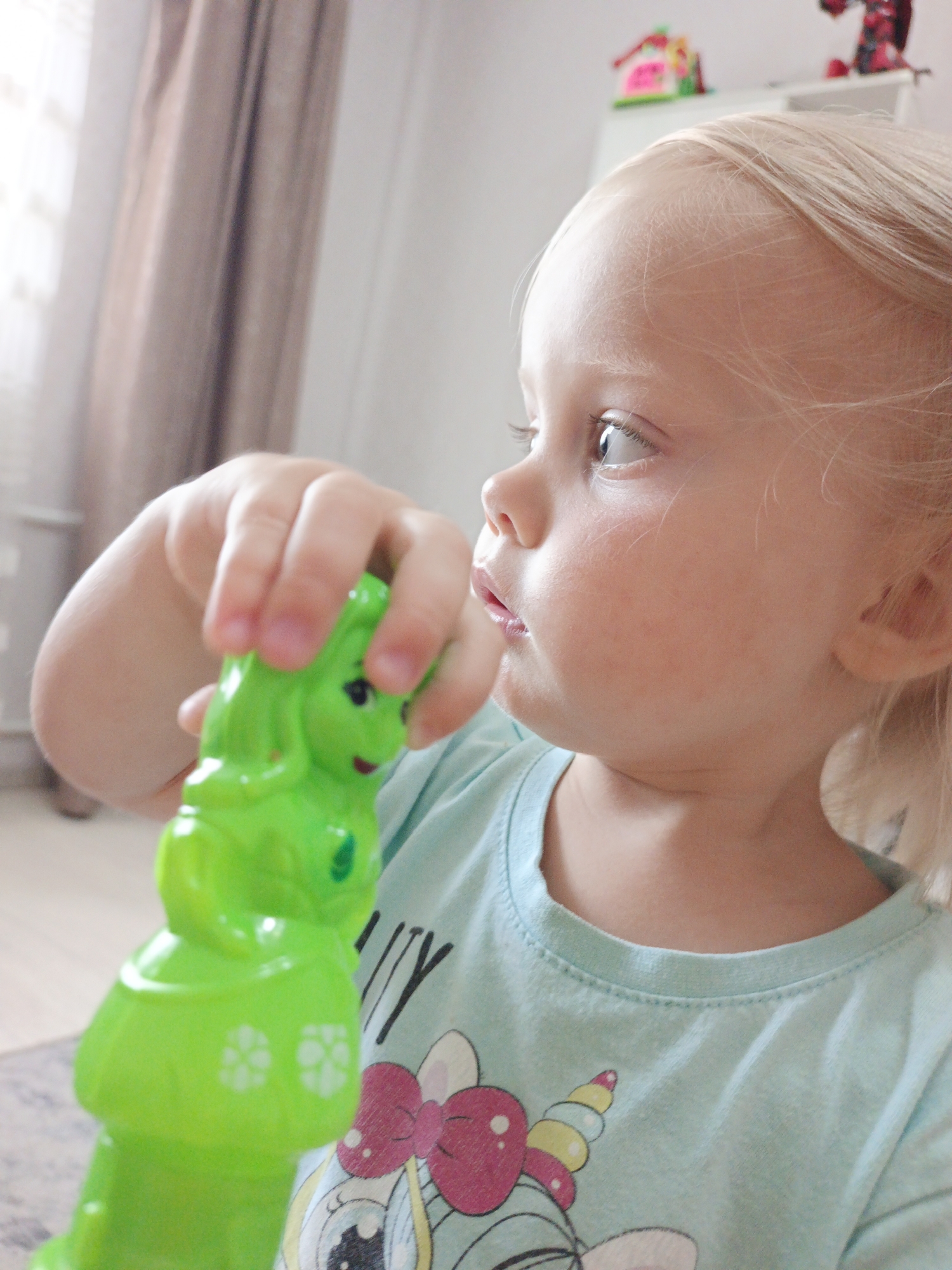 A young child, likely a girl with blonde hair, is shown in profile, looking to the left. She is wearing a light blue t-shirt with a unicorn graphic. Her right hand is holding a bright green plastic toy, possibly a stacking toy or a figure. The background is slightly out of focus, showing a beige curtain and what appears to be a shelf with colorful toys in the distance. The lighting suggests an indoor setting, possibly a home. No specific location in Zlatoust, Russia can be determined from the image.