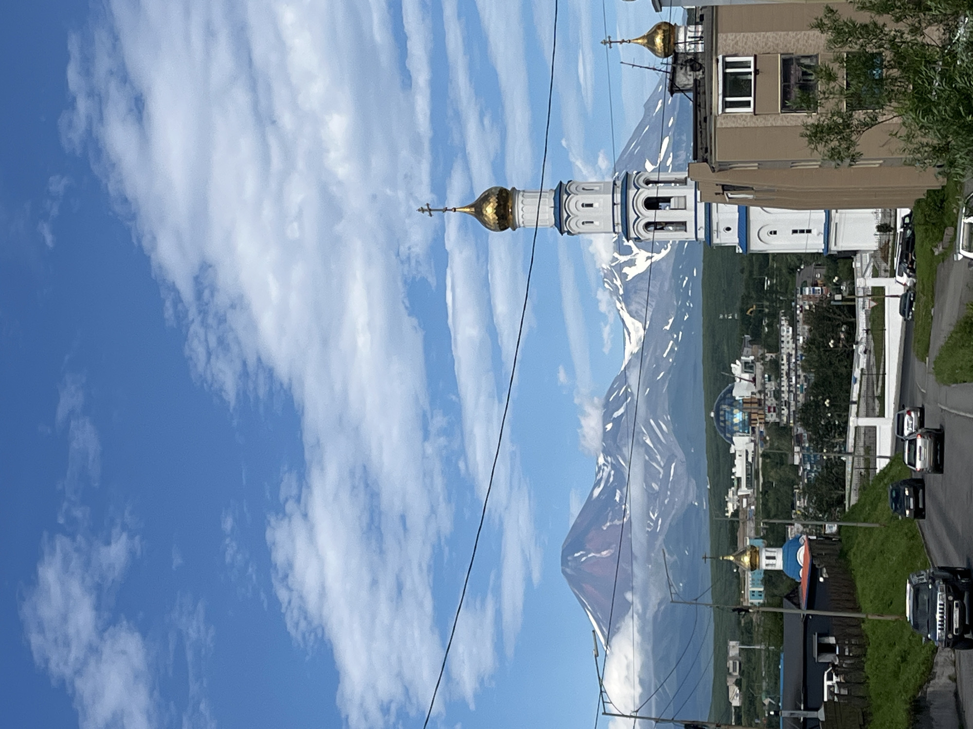 The image depicts a daytime outdoor scene in the city of Petropavlovsk-Kamchatsky, Russia. In the foreground, a two-lane road curves uphill with several vehicles driving on it. To the right of the road, a multi-story building with a beige facade is visible, and to the left, a grassy slope borders the road.
Dominating the mid-ground are several church structures. A prominent white bell tower with a golden dome and a cross stands tall, adorned with blue accents. Another smaller golden dome with a cross is also visible on a separate structure. Behind these, a cityscape unfolds with various buildings and structures, including a dome-shaped building.
The background features a majestic mountain range, partially covered in snow, under a bright blue sky with scattered white clouds. The mountains appear to be volcanoes, a characteristic feature of the Kamchatka Peninsula. Power lines are visible in the foreground and mid-ground, crossing the frame horizontally. The overall impression is a blend of urban and natural landscapes, with religious architecture playing a significant role in the foreground and mid-ground.