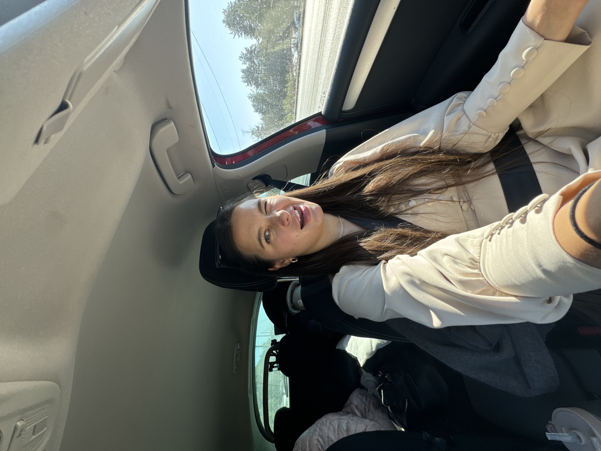 A young woman is in the back seat of a car, possibly a passenger. She has long, dark hair and is wearing a light-colored jacket. She is sticking her tongue out and winking, suggesting she is taking a selfie. The car is moving; trees and a road can be seen through the car's window. The interior of the car is visible, including the headrest of the front seat, and some items in the back. No identifiable information is in the image that confirms the location is Vladimir, Russia.