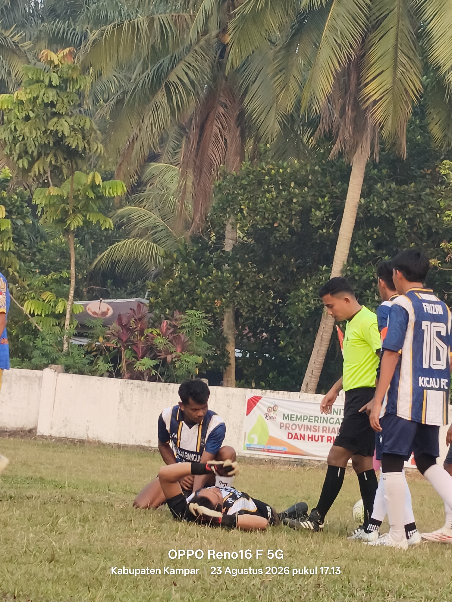 In Penghidupan, Indonesia, two young males are engaged in a soccer game. One player, wearing a dark and white jersey with the number 16 and the text "KICAU FC," lies on the grass, seemingly injured. Another player, dressed in a neon green shirt and black shorts, stands over him, possibly tending to the injured player. A third male, also in a dark and white jersey, stands behind them, watching the situation. The game takes place on a grass field, with palm trees and lush green foliage forming the background, suggesting a tropical environment. A white fence runs along the edge of the field, and a banner with Indonesian text is visible behind the players. The scene is captured outdoors during daylight hours.